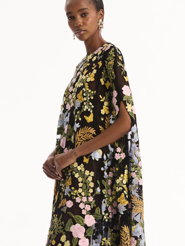 The model is wearing the Dominican Mixed-Floral Guipure Caftan. Featuring a long, flowing silhouette draping from the shoulders. Detail View.