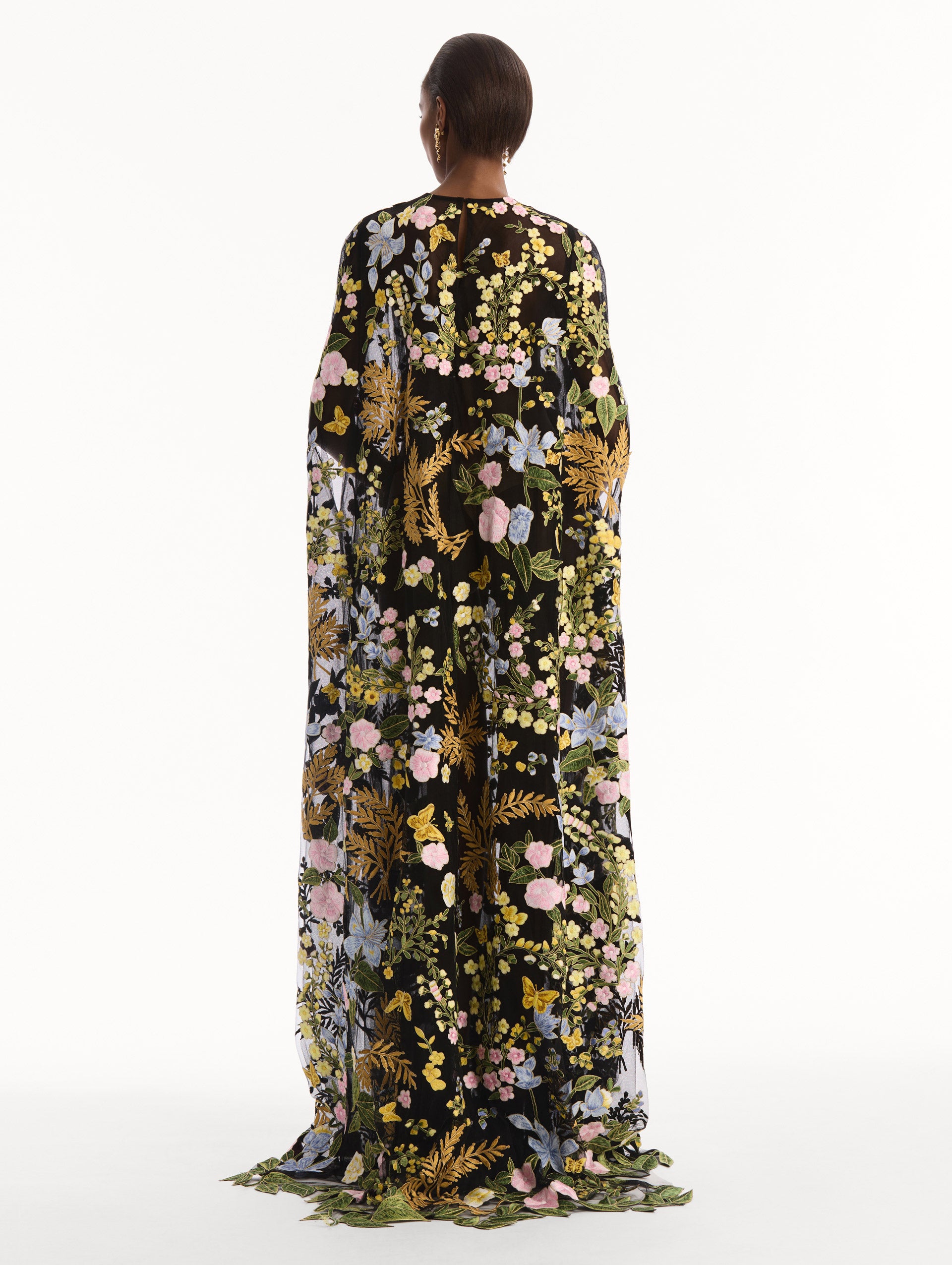 The model is wearing the Dominican Mixed-Floral Guipure Caftan. Featuring a long, flowing silhouette draping from the shoulders. Back View.