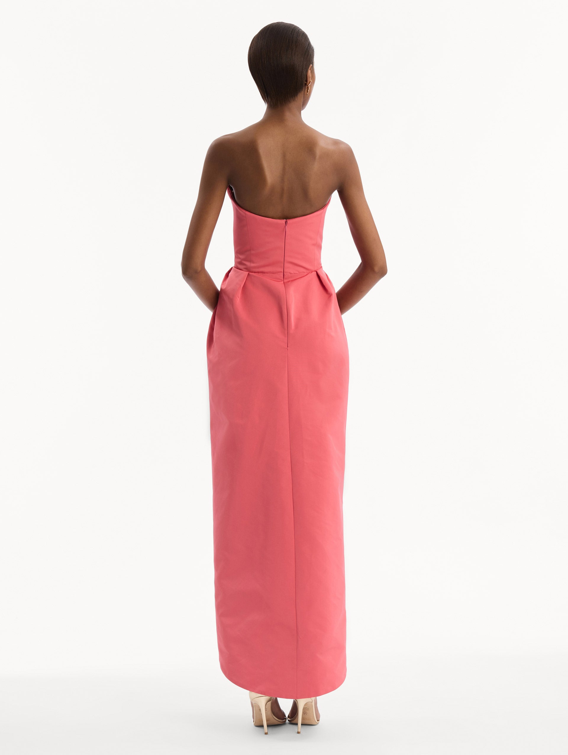 The model is wearing the Strapless Faille Gown in Guava Pink. Featuring a sculpted silhouette and wrap-style skirt with an asymmetrical hem. Back View.