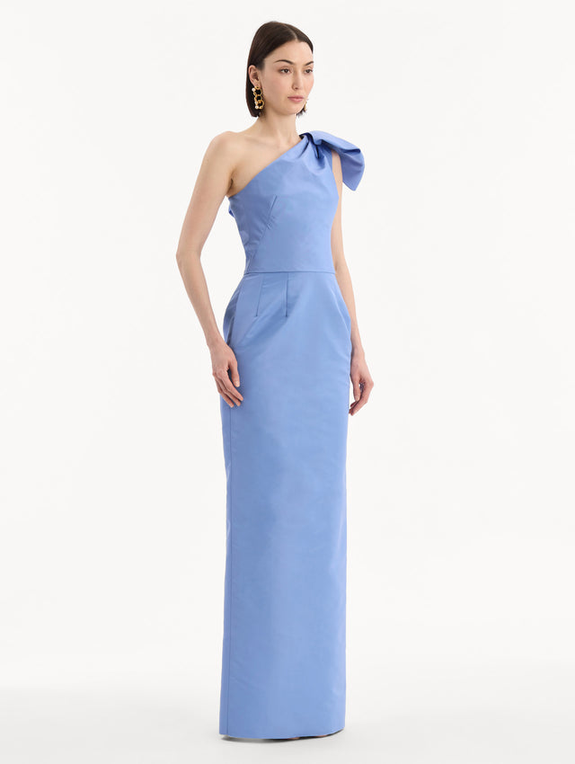 The model is wearing the Bow-Detail Faille Column Gown in a periwinkle blue. Featuring a column silhouette and sculptural bow detail at the shoulder. Side View.