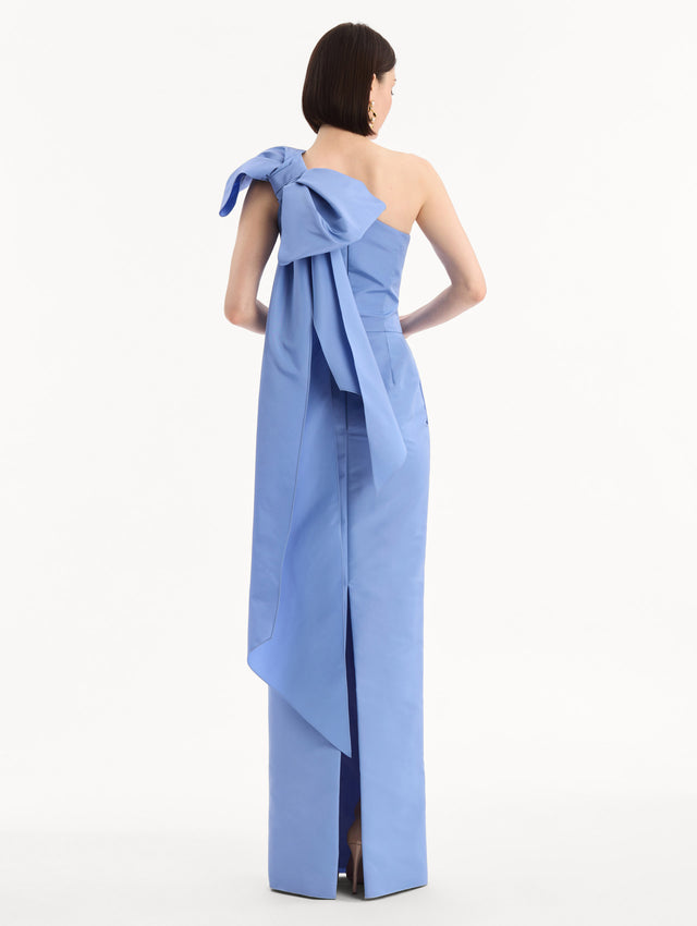 The model is wearing the Bow-Detail Faille Column Gown in a periwinkle blue. Featuring a column silhouette and sculptural bow detail at the shoulder. Back View.