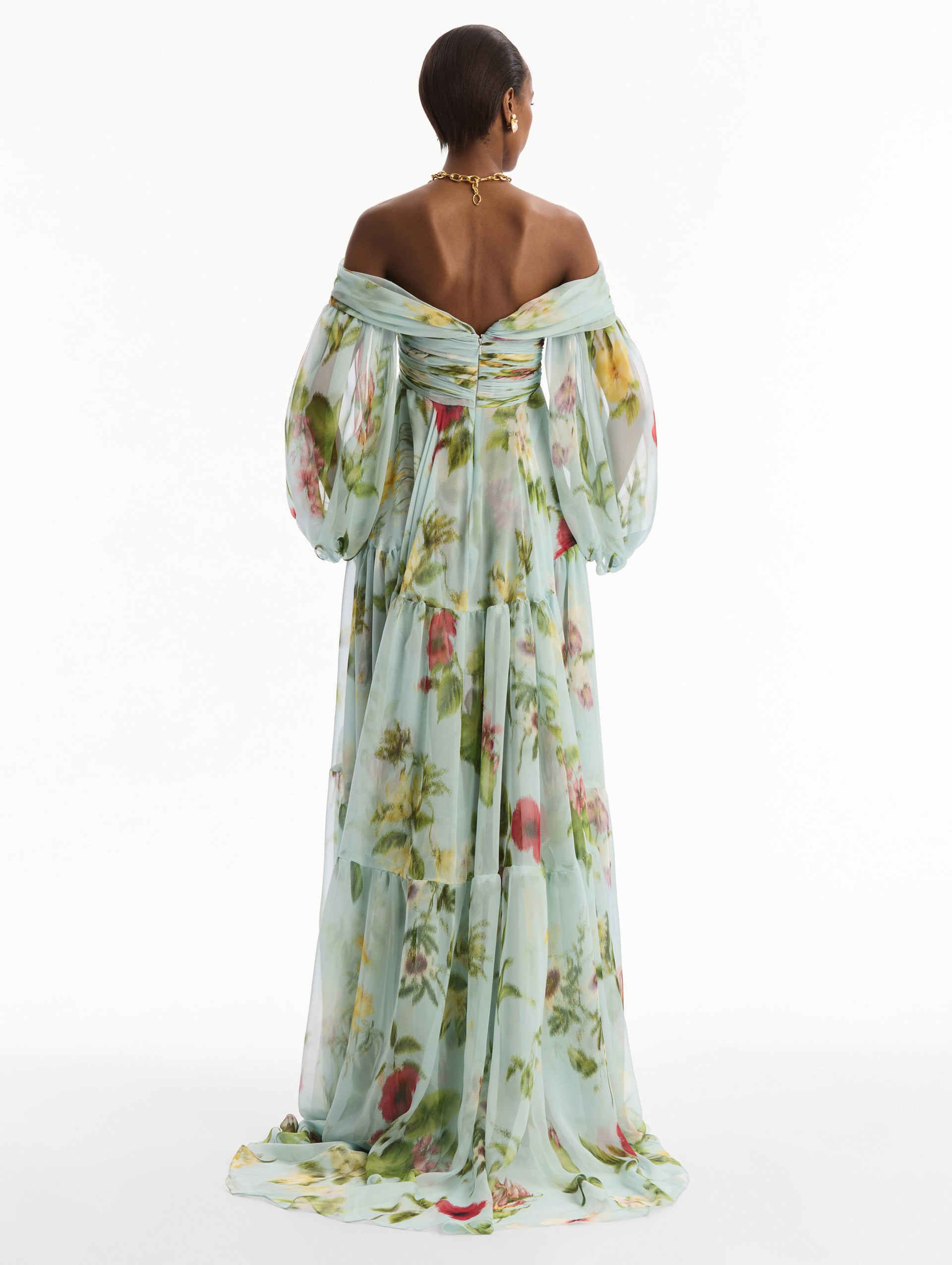 The model is wearing the Chiné Dominican Mixed-Floral Chiffon Sweetheart Gown. Featuring an off-the-shoulder sweetheart neckline, long sleeves, and flowing, tiered skirt. Back View.