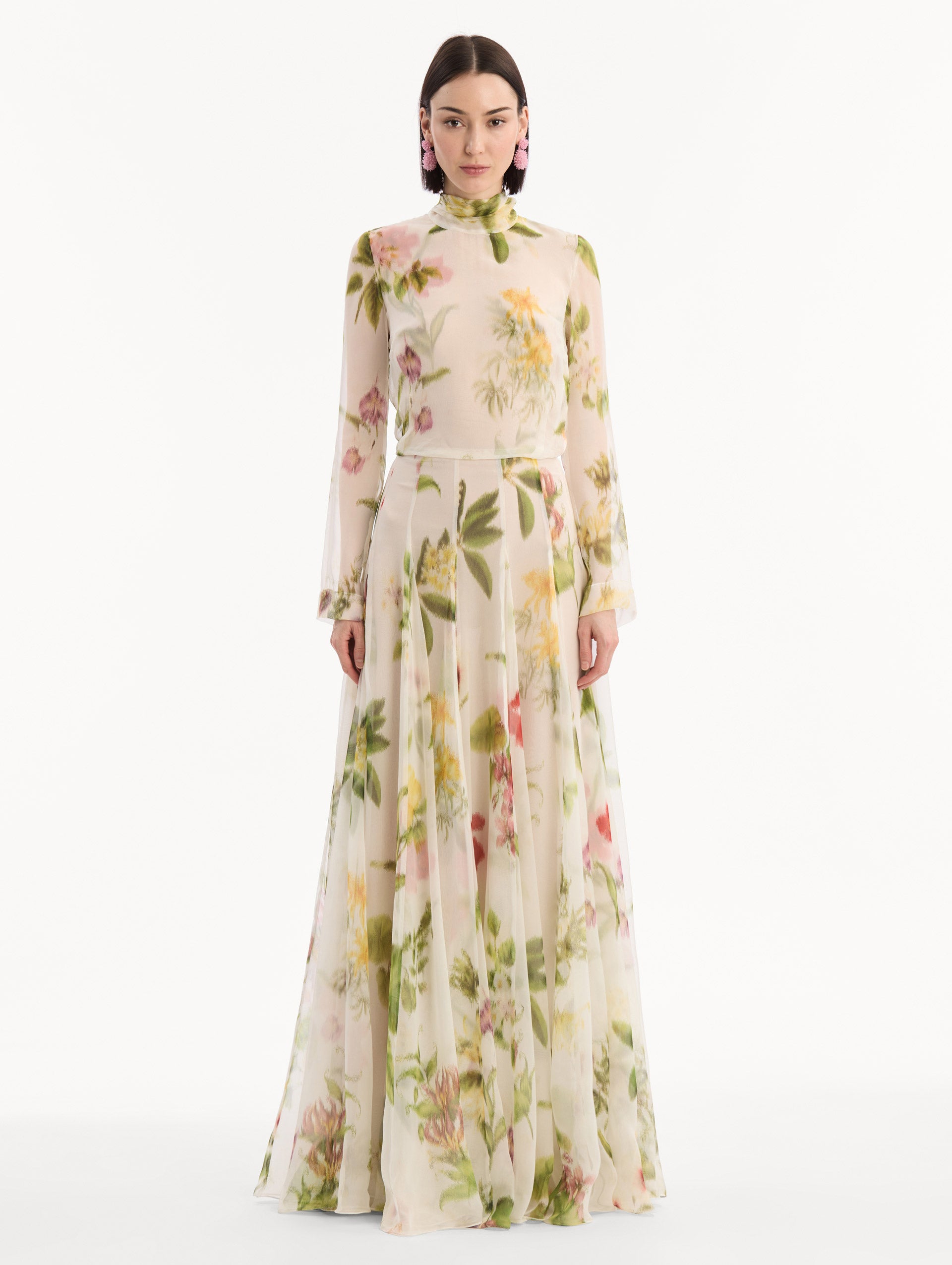 The model is wearing the Chiné Dominican Mixed-Floral Chiffon Gown in Ivory printed with an array of Dominican florals. Featuring long sleeves, mock neck, and flowing, pleated skirt. Front View.