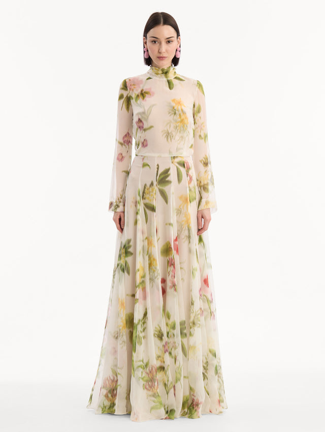 The model is wearing the Chiné Dominican Mixed-Floral Chiffon Gown in Ivory printed with an array of Dominican florals. Featuring long sleeves, mock neck, and flowing, pleated skirt. Front View.