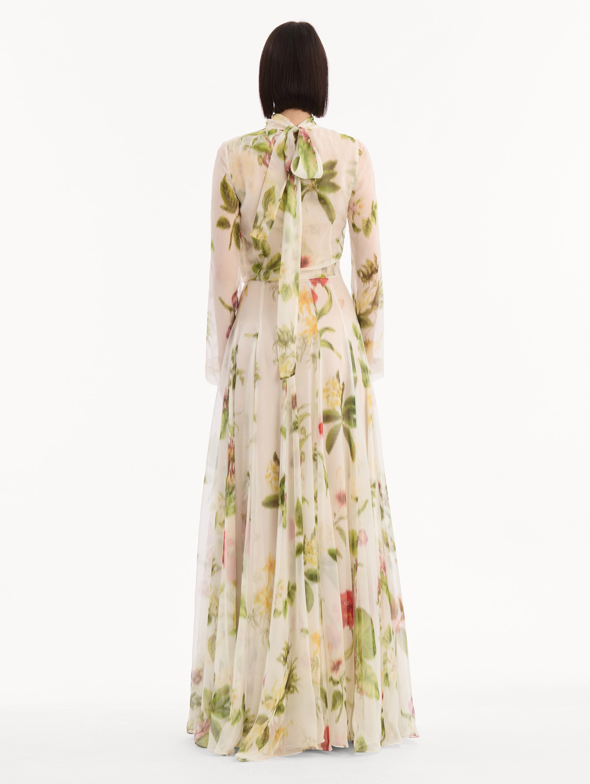 The model is wearing the Chiné Dominican Mixed-Floral Chiffon Gown in Ivory printed with an array of Dominican florals. Featuring long sleeves, mock neck, and flowing, pleated skirt. Back View.