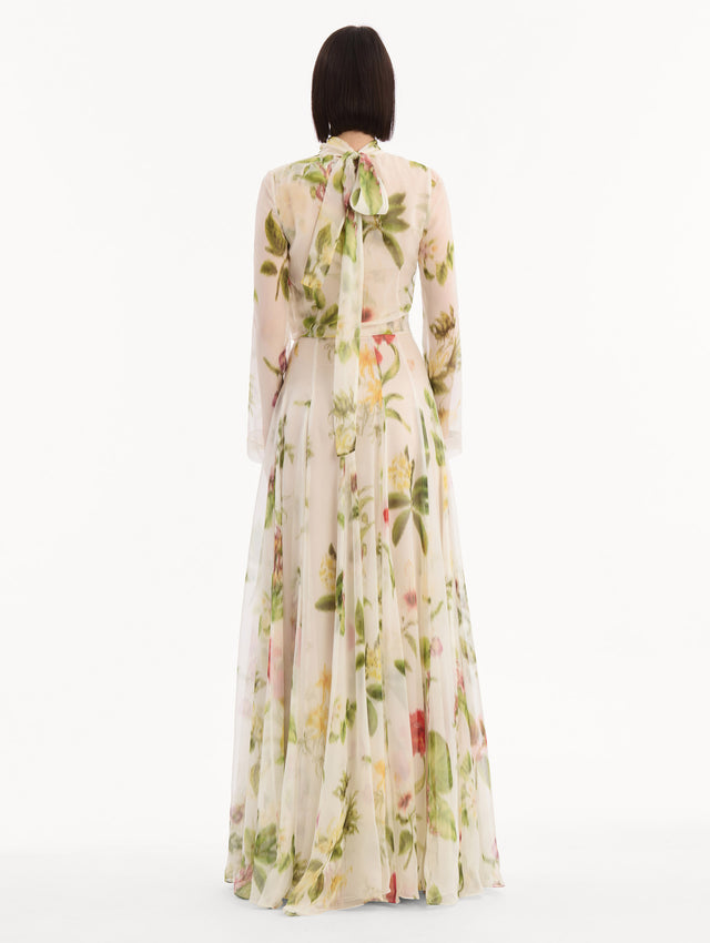 The model is wearing the Chiné Dominican Mixed-Floral Chiffon Gown in Ivory printed with an array of Dominican florals. Featuring long sleeves, mock neck, and flowing, pleated skirt. Back View.