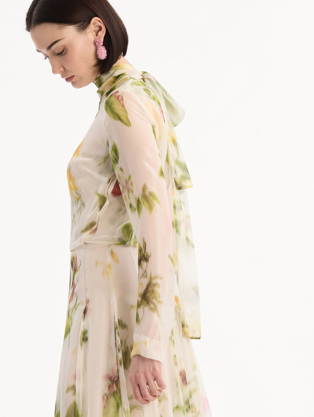 The model is wearing the Chiné Dominican Mixed-Floral Chiffon Gown in Ivory printed with an array of Dominican florals. Featuring long sleeves, mock neck, and flowing, pleated skirt. Detail View.