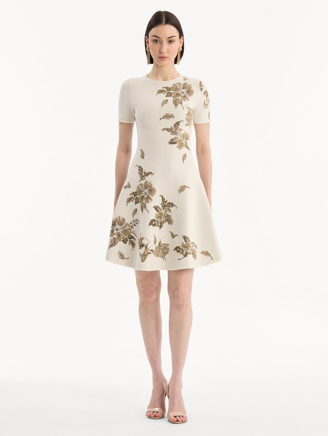 The model is wearing the Hibiscus Metallic Jacquard Dress in Ivory with Gold hibiscus motifs. Featuring short sleeves, a softly structured silhouette, and mini-length skirt. Front View.