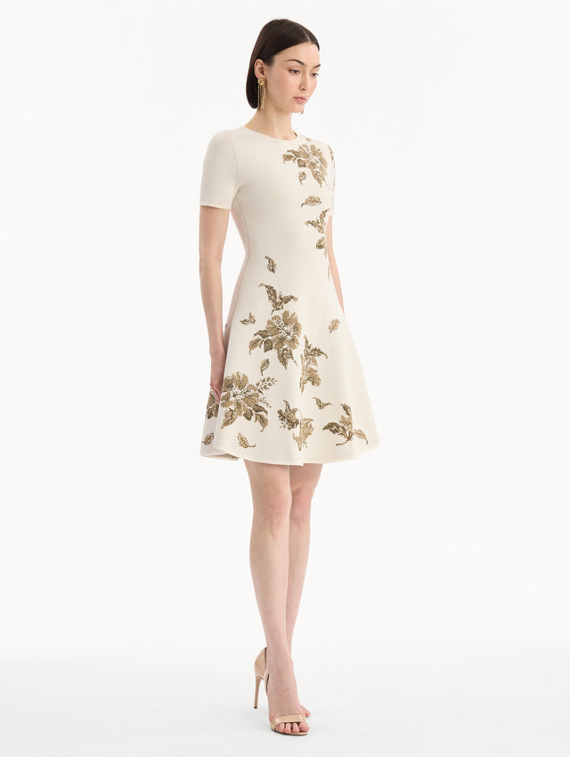 The model is wearing the Hibiscus Metallic Jacquard Dress in Ivory with Gold hibiscus motifs. Featuring short sleeves, a softly structured silhouette, and mini-length skirt. Side View.
