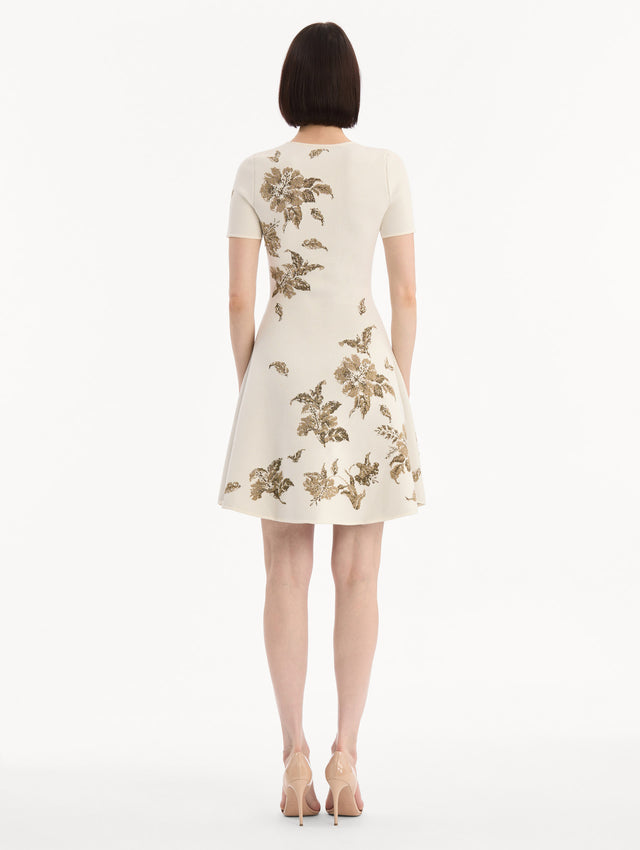 The model is wearing the Hibiscus Metallic Jacquard Dress in Ivory with Gold hibiscus motifs. Featuring short sleeves, a softly structured silhouette, and mini-length skirt. Back View.