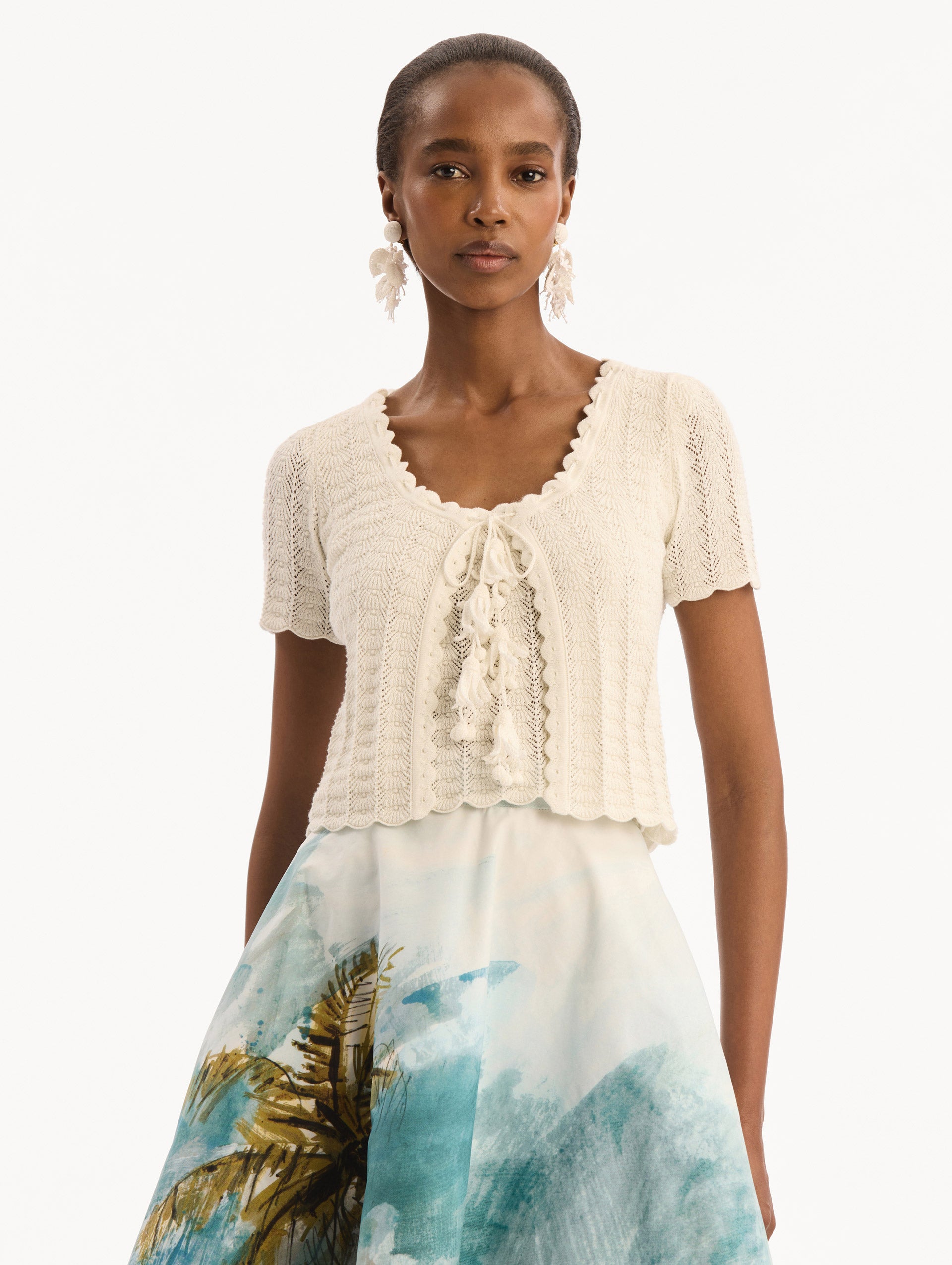 The model is wearing the Pointelle Weave Cardigan in Ivory. Featuring a scoop neckline, short sleeves, scalloped edges, and a front tie closure. Front View.