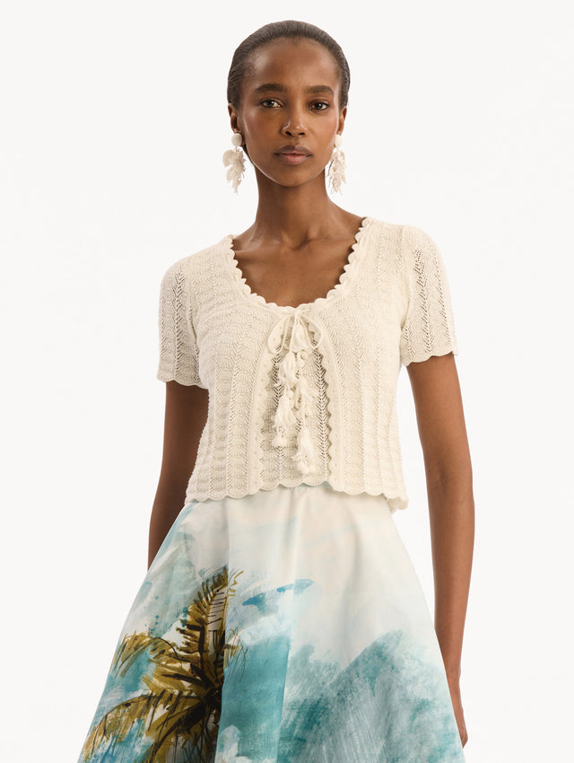 The model is wearing the Pointelle Weave Cardigan in Ivory. Featuring a scoop neckline, short sleeves, scalloped edges, and a front tie closure. Front View.