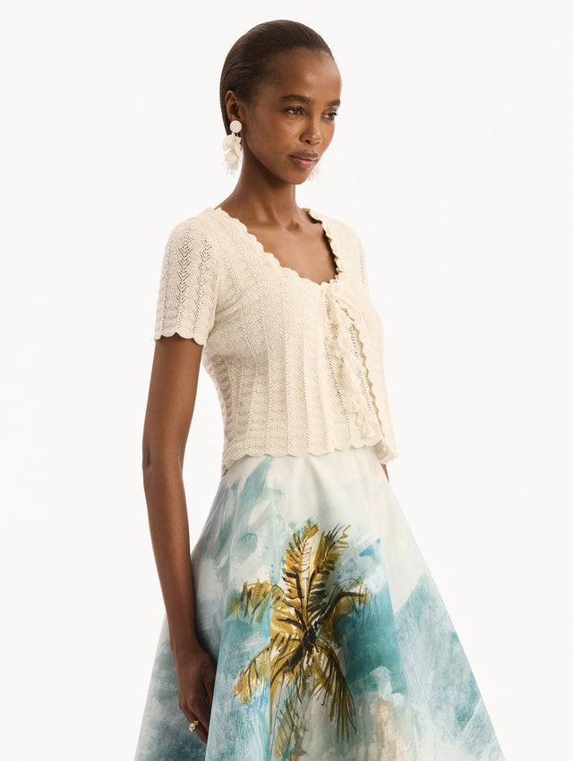 The model is wearing the Pointelle Weave Cardigan in Ivory. Featuring a scoop neckline, short sleeves, scalloped edges, and a front tie closure. Side View.