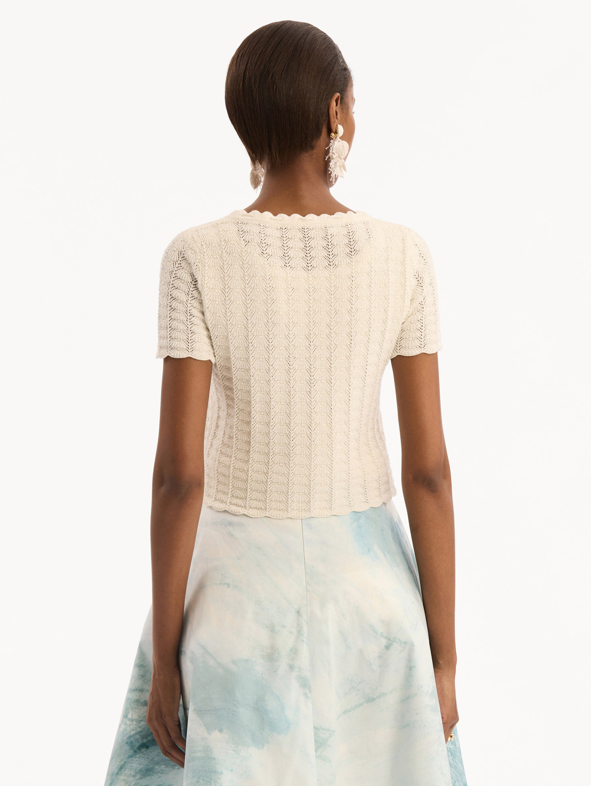 The model is wearing the Pointelle Weave Cardigan in Ivory. Featuring a scoop neckline, short sleeves, scalloped edges, and a front tie closure. Back View.