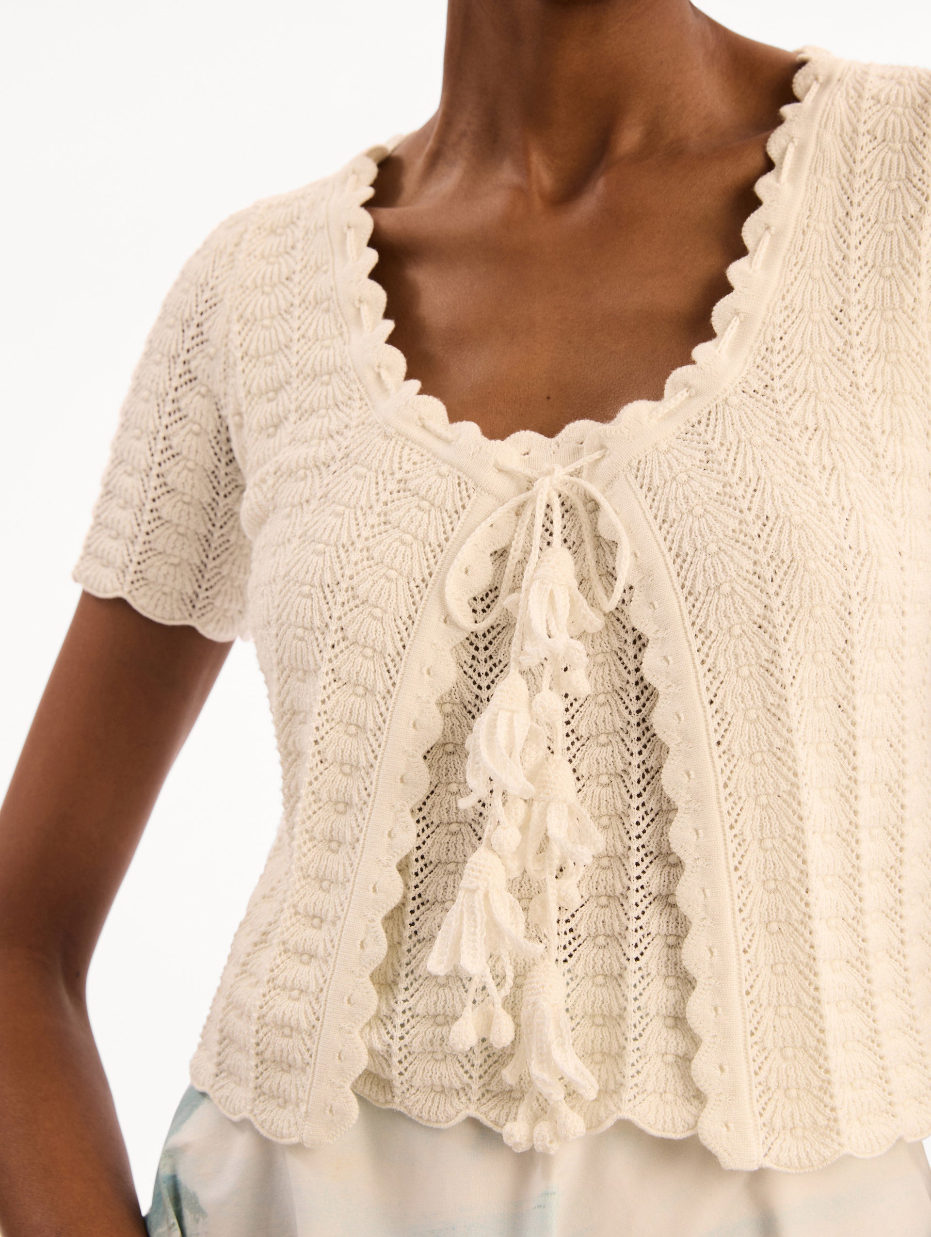 The model is wearing the Pointelle Weave Cardigan in Ivory. Featuring a scoop neckline, short sleeves, scalloped edges, and a front tie closure. Detail View.
