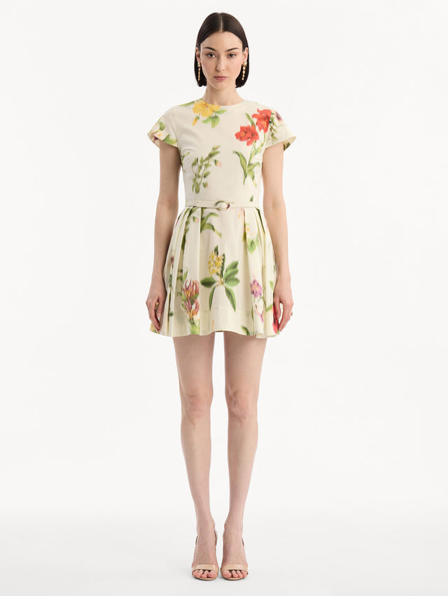 The model is wearing the Chiné Dominican Mixed-Floral Cotton Poplin Dress in Ivory printed with an array of Dominican florals. Featuring cap sleeves, belt at waist, and gathered mini skirt. Front View.