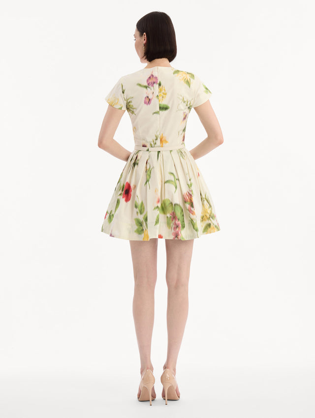 The model is wearing the Chiné Dominican Mixed-Floral Cotton Poplin Dress in Ivory printed with an array of Dominican florals. Featuring cap sleeves, belt at waist, and gathered mini skirt. Back View.
