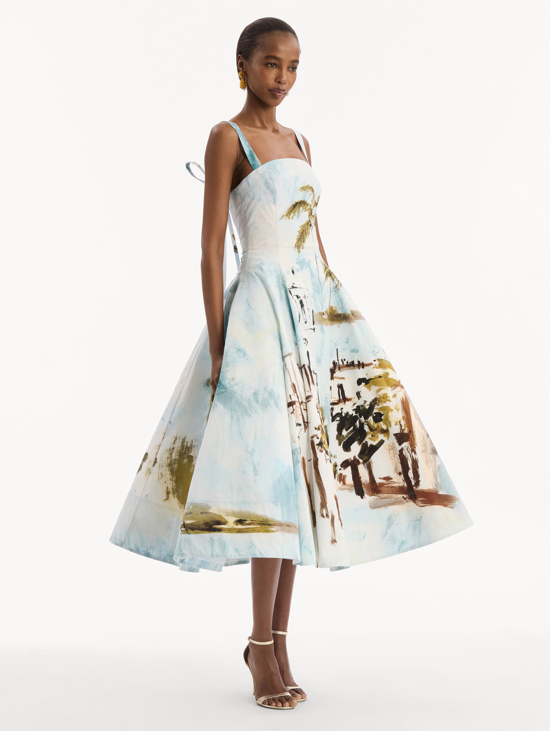 The model is wearing the Dominican Landscape Cotton Poplin Sleeveless Dress. Featuring tank straps with bow feature in back, and a full, midi-length skirt. Side View.