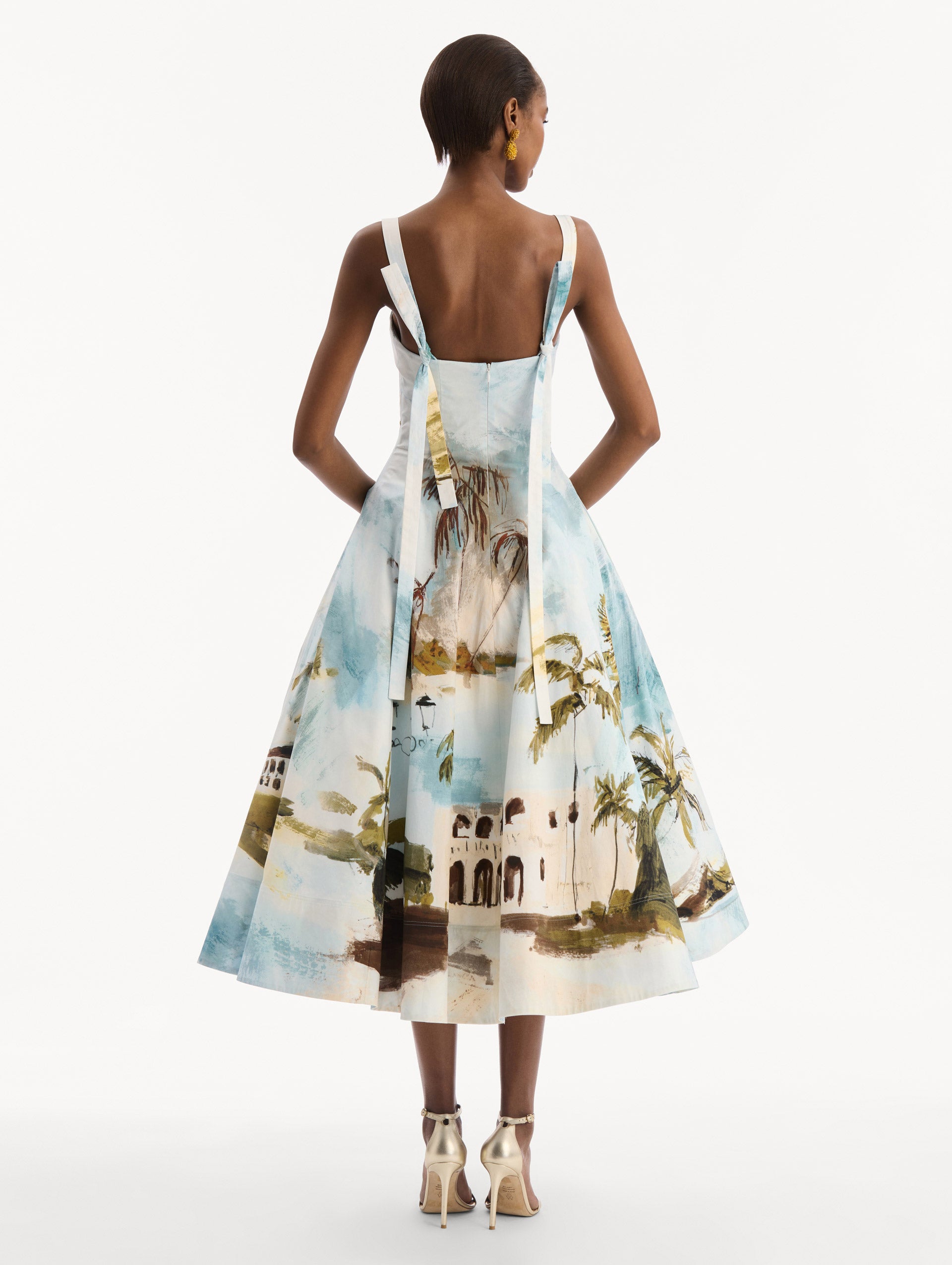 The model is wearing the Dominican Landscape Cotton Poplin Sleeveless Dress. Featuring tank straps with bow feature in back, and a full, midi-length skirt. Back View.