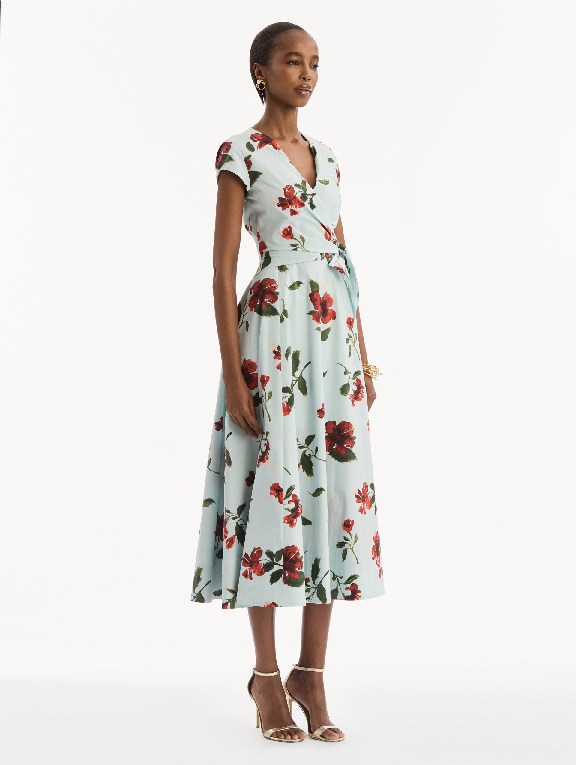 The model is wearing the Hibiscus Cotton Poplin Wrap Dress in light blue printed with red hibiscus blooms. Featuring a wrap construction bodice, cap sleeves, V-neckline, and full midi skirt. Side View.