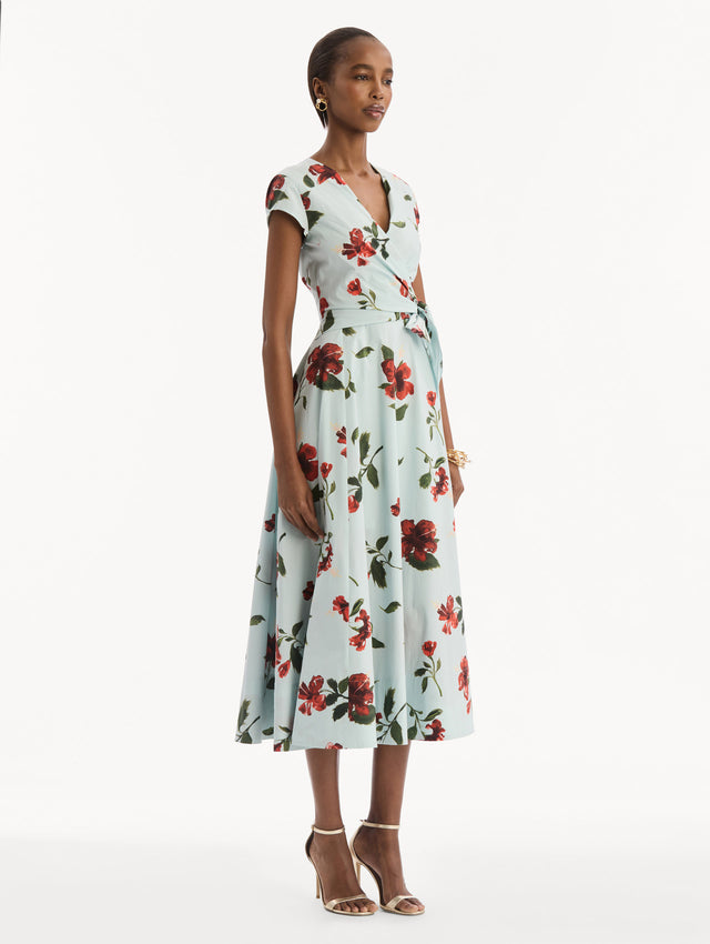 The model is wearing the Hibiscus Cotton Poplin Wrap Dress in light blue printed with red hibiscus blooms. Featuring a wrap construction bodice, cap sleeves, V-neckline, and full midi skirt. Side View.
