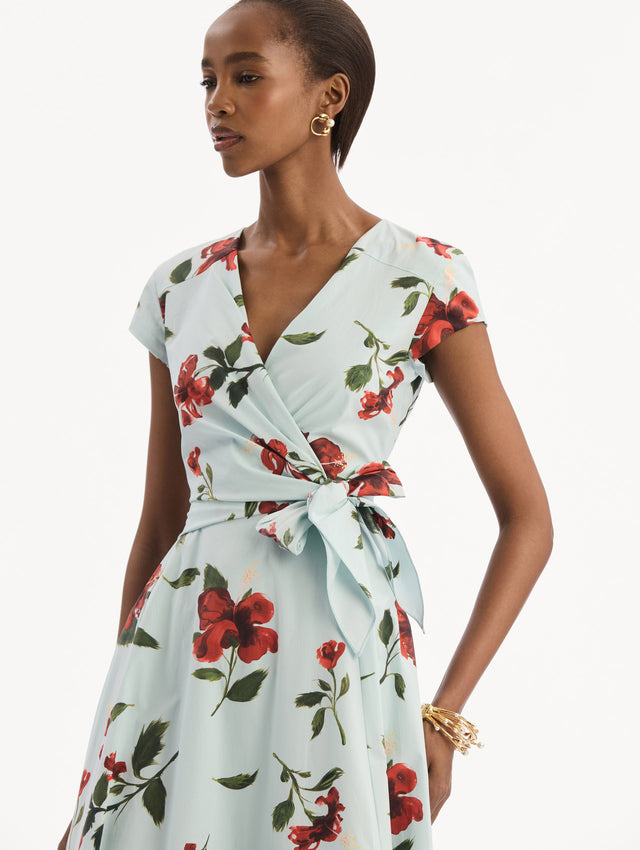 The model is wearing the Hibiscus Cotton Poplin Wrap Dress in light blue printed with red hibiscus blooms. Featuring a wrap construction bodice, cap sleeves, V-neckline, and full midi skirt. Detail View.