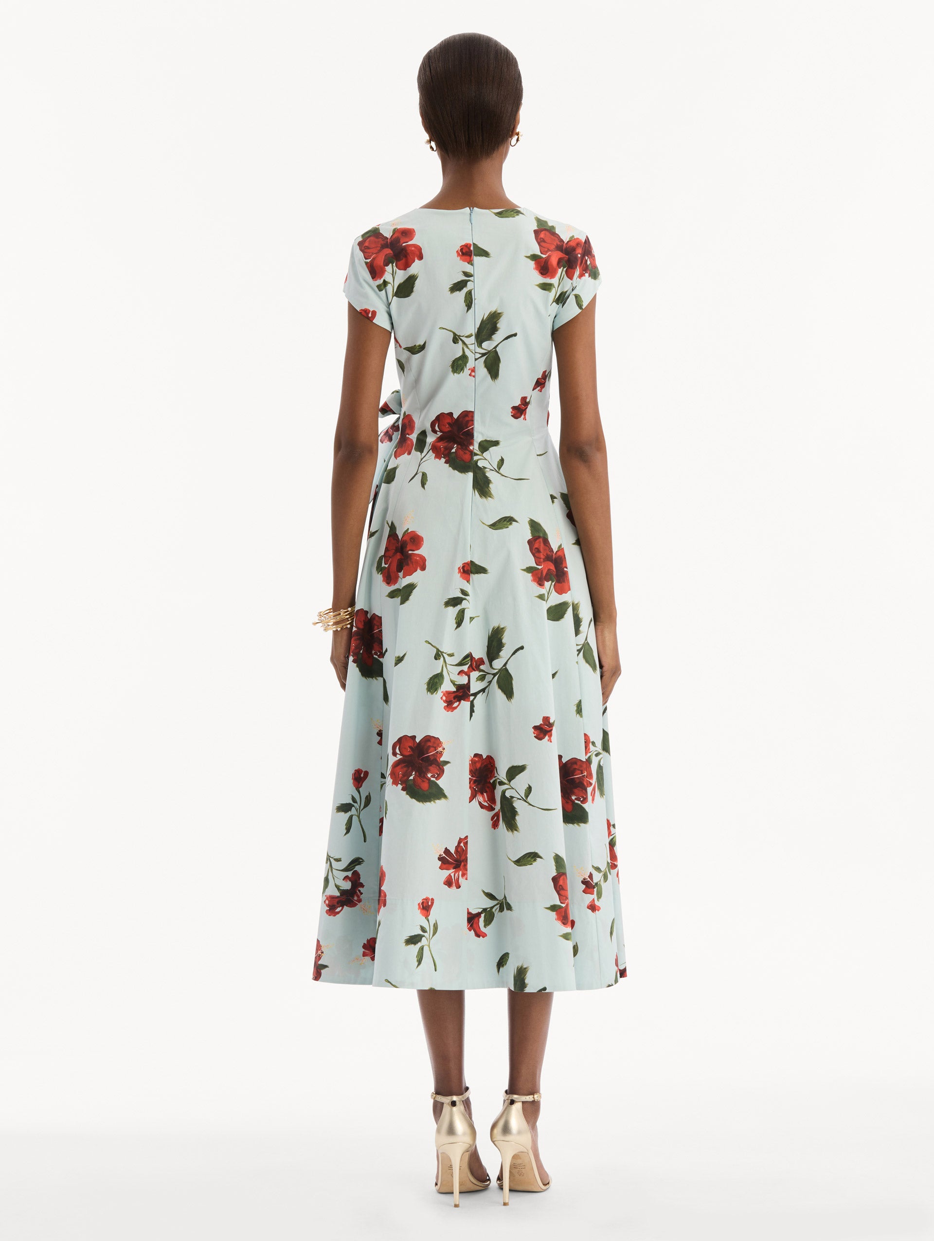 The model is wearing the Hibiscus Cotton Poplin Wrap Dress in light blue printed with red hibiscus blooms. Featuring a wrap construction bodice, cap sleeves, V-neckline, and full midi skirt. Back View.