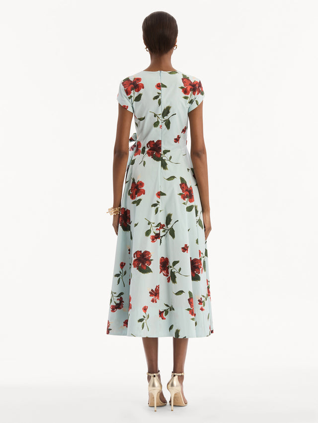 The model is wearing the Hibiscus Cotton Poplin Wrap Dress in light blue printed with red hibiscus blooms. Featuring a wrap construction bodice, cap sleeves, V-neckline, and full midi skirt. Back View.