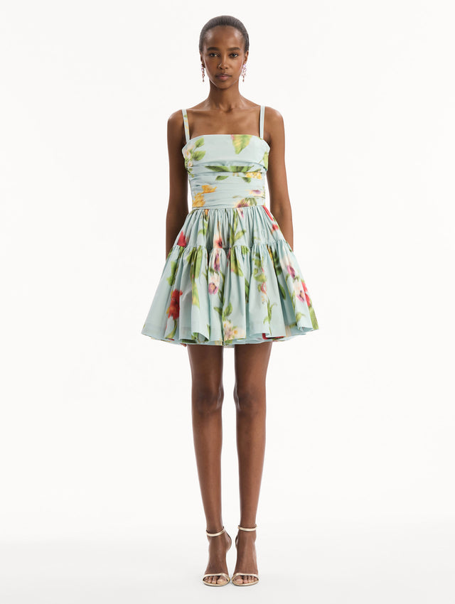 The model is wearing the Chiné Dominican Mixed-Floral Cotton Poplin Tiered Dress in light blue printed with a vibrant array of Dominican florals. Featuring a ruched bodice, tank straps, and gathered mini skirt. Front View.