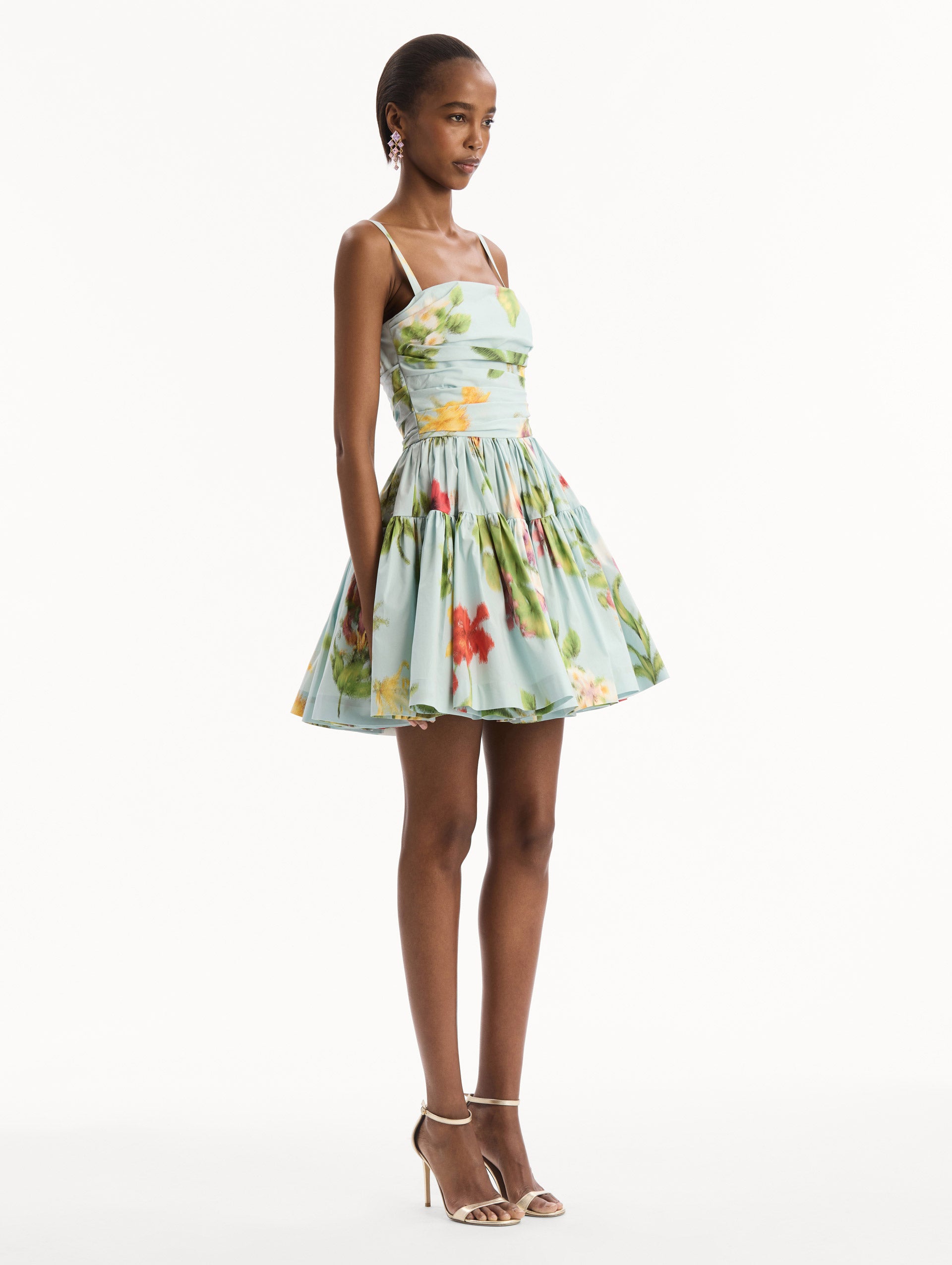 The model is wearing the Chiné Dominican Mixed-Floral Cotton Poplin Tiered Dress in light blue printed with a vibrant array of Dominican florals. Featuring a ruched bodice, tank straps, and gathered mini skirt. Side View.