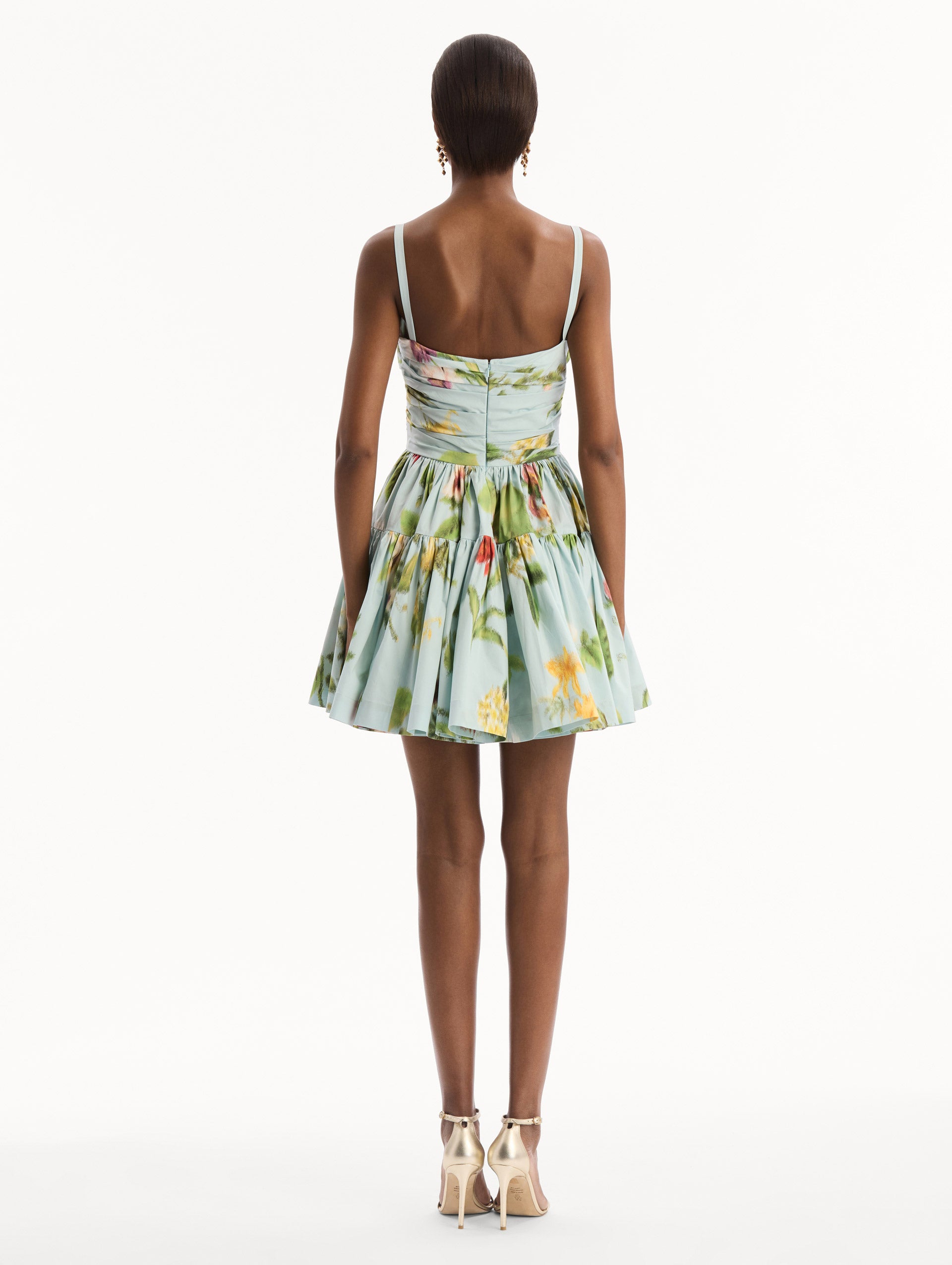 The model is wearing the Chiné Dominican Mixed-Floral Cotton Poplin Tiered Dress in light blue printed with a vibrant array of Dominican florals. Featuring a ruched bodice, tank straps, and gathered mini skirt. Back View.
