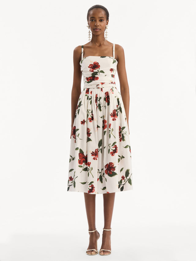 The model is wearing the Hibiscus Cotton Poplin Dress in white with red hibiscus blooms. Featuring a ruched bodice, tank straps, belt at waist, and full, gathered skirt. Front View.
