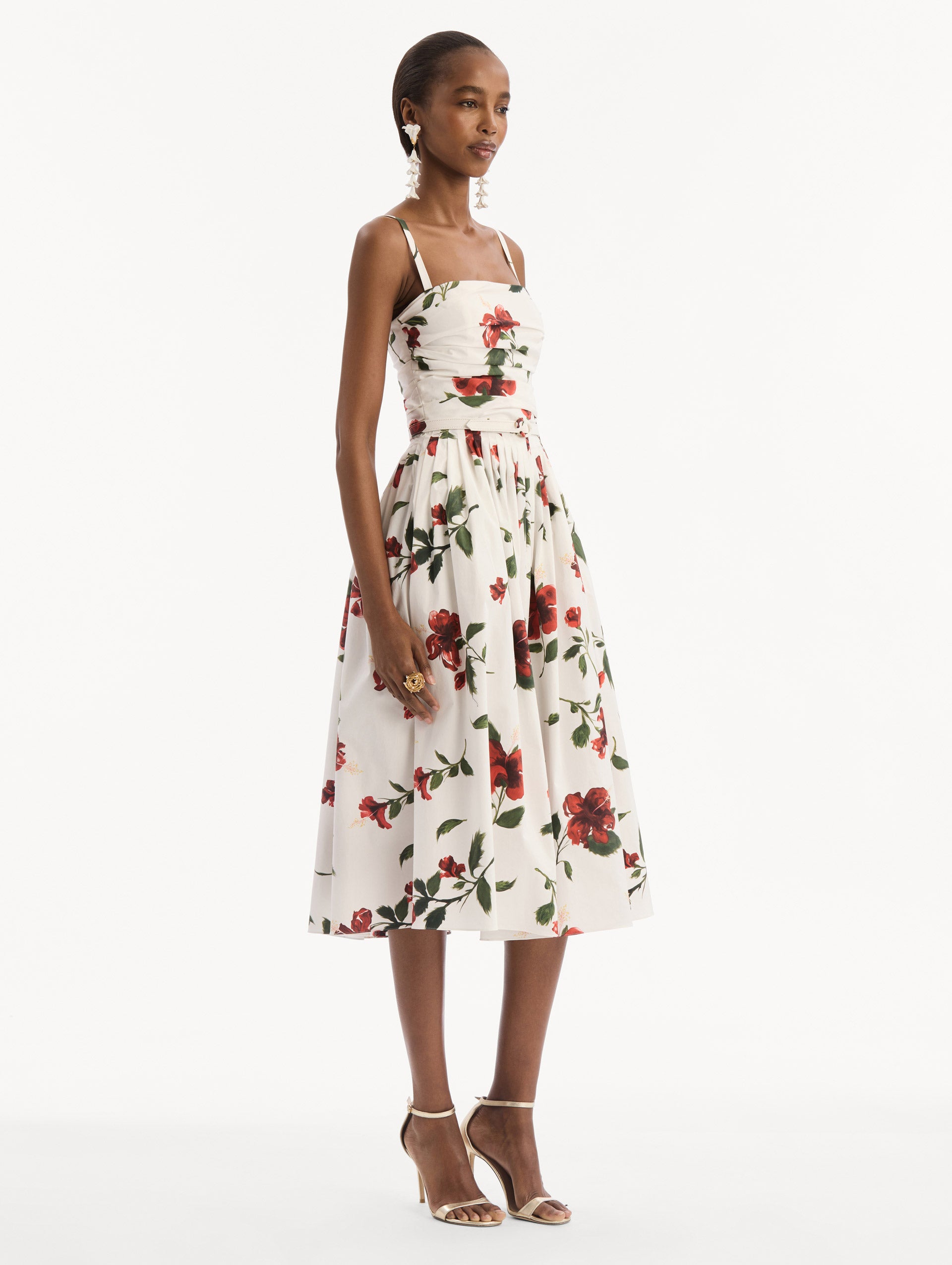 The model is wearing the Hibiscus Cotton Poplin Dress in white with red hibiscus blooms. Featuring a ruched bodice, tank straps, belt at waist, and full, gathered skirt. Side View.