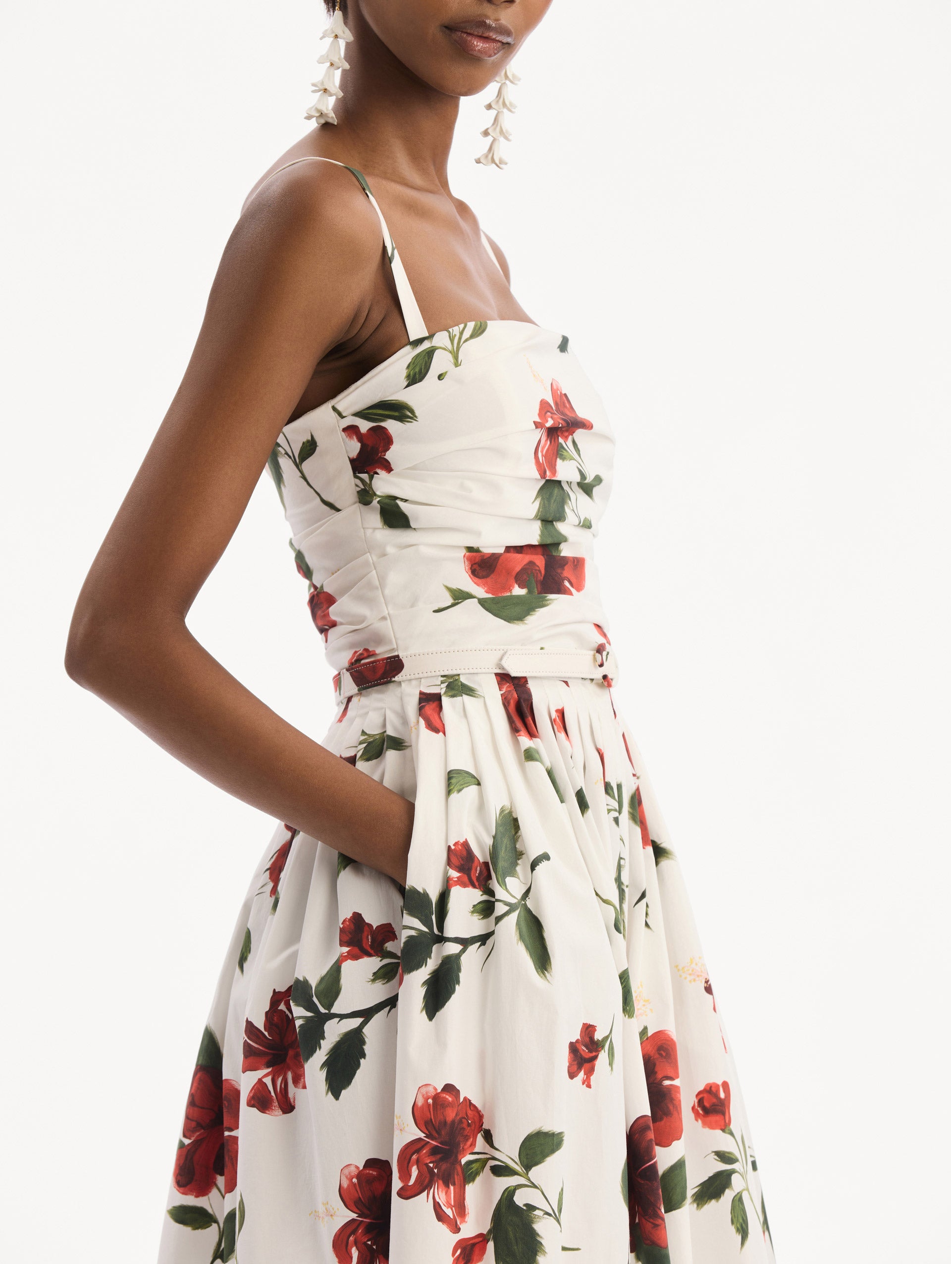 The model is wearing the Hibiscus Cotton Poplin Dress in white with red hibiscus blooms. Featuring a ruched bodice, tank straps, belt at waist, and full, gathered skirt. Detail View.