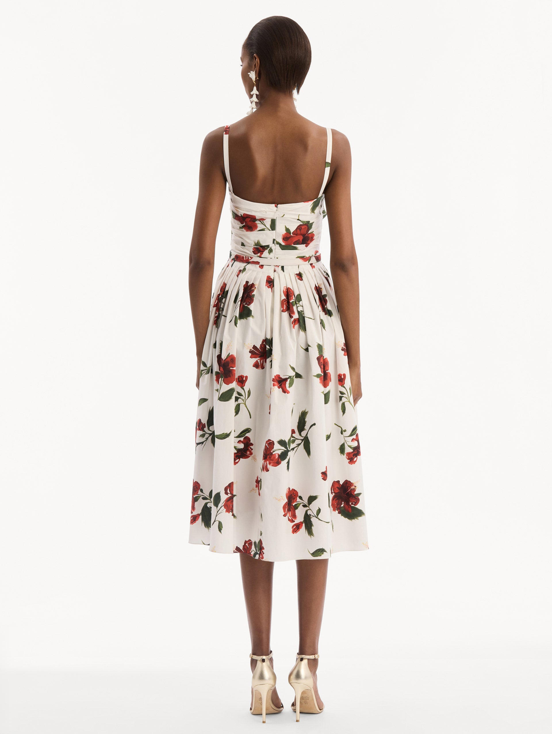The model is wearing the Hibiscus Cotton Poplin Dress in white with red hibiscus blooms. Featuring a ruched bodice, tank straps, belt at waist, and full, gathered skirt. Back View.