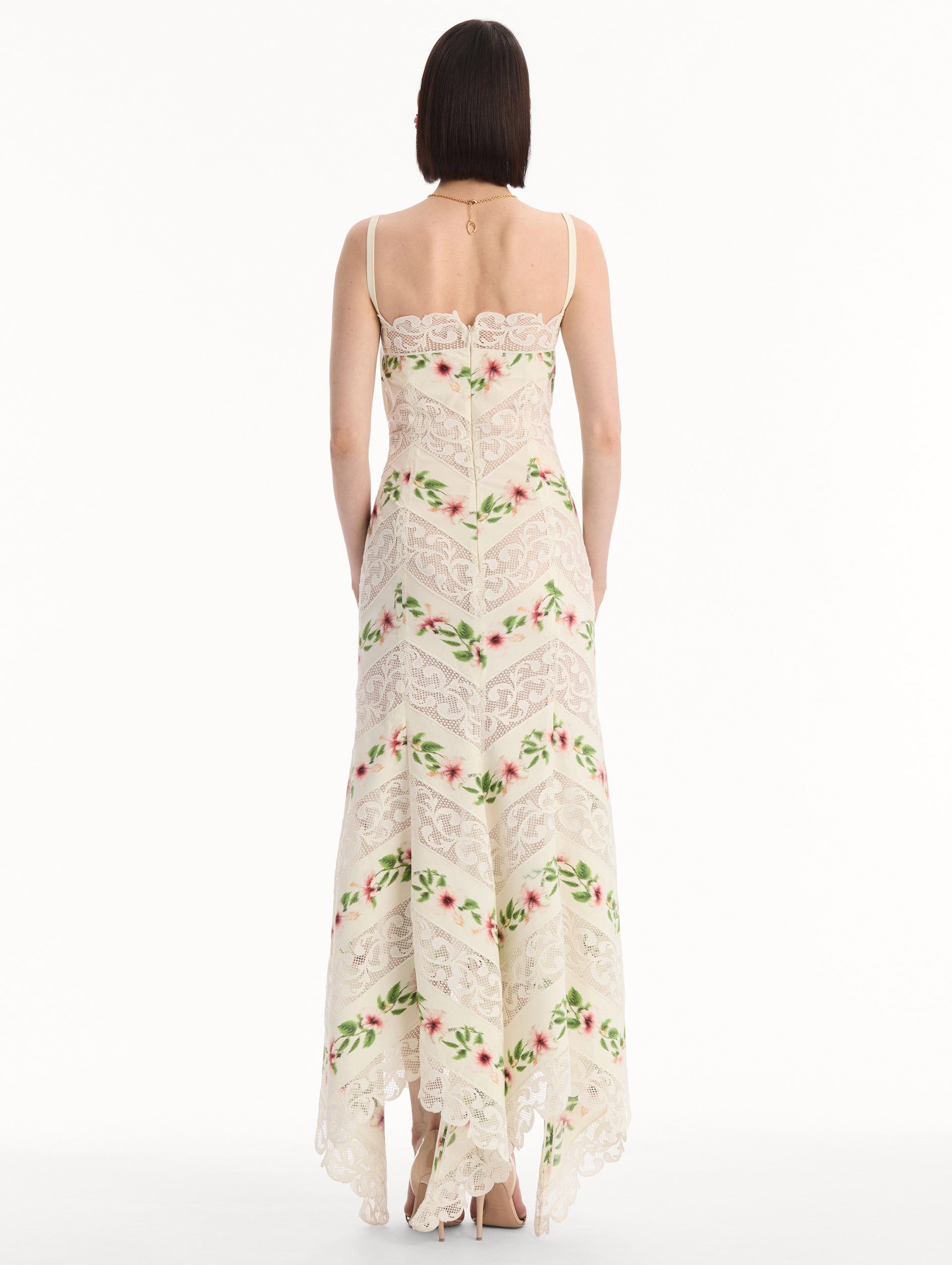 The model is wearing the Chiné Hibiscus Striped Cotton Poplin Dress in Ivory. Featuring a lace neckline, tank straps, and woven hibiscus motifs alternating with panels of faille and guipure lace, creating a striped composition. Back View.