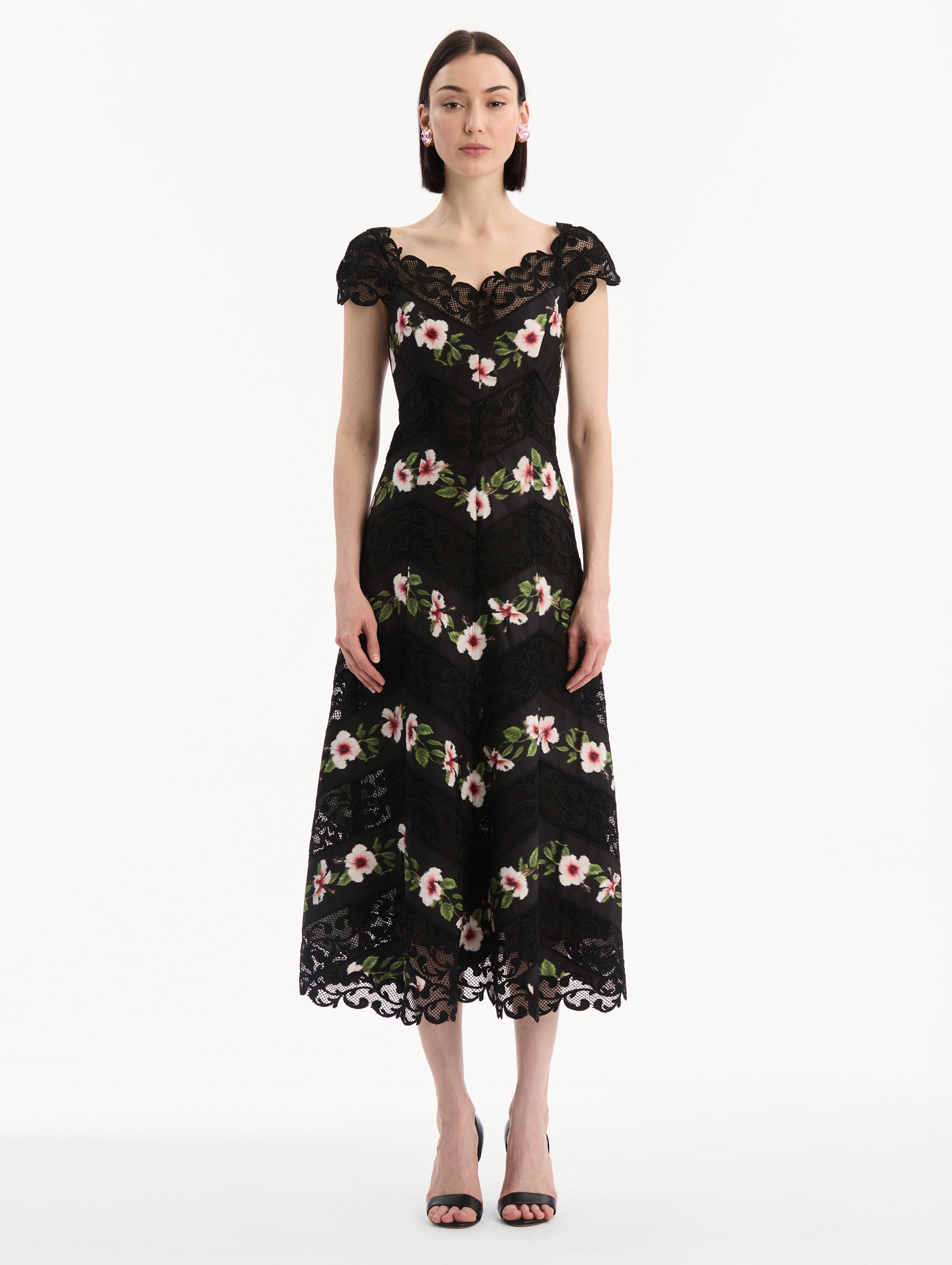 The model is wearing the Chiné Hibiscus Striped Faille Midi Dress in Black. Featuring off-the-shoulder cap sleeves, a midi length, and woven hibiscus motifs alternating with panels of faille and guipure lace, creating a striped composition. Front View.