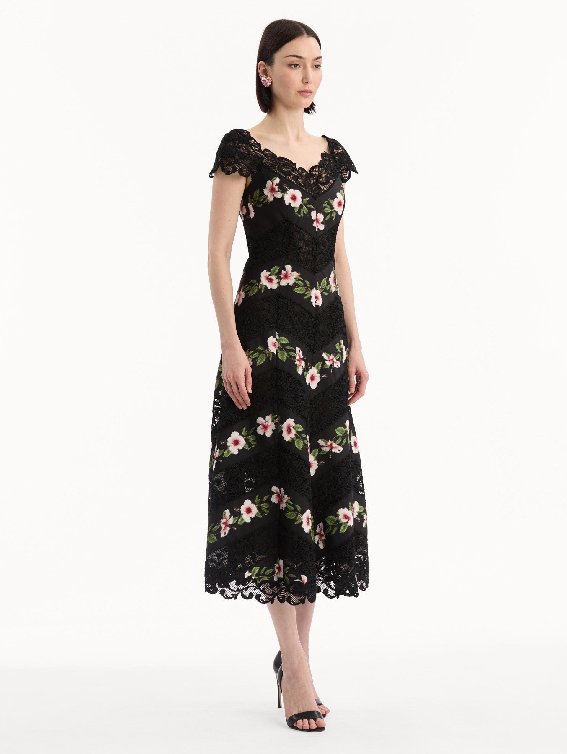 The model is wearing the Chiné Hibiscus Striped Faille Midi Dress in Black. Featuring off-the-shoulder cap sleeves, a midi length, and woven hibiscus motifs alternating with panels of faille and guipure lace, creating a striped composition. Side View.