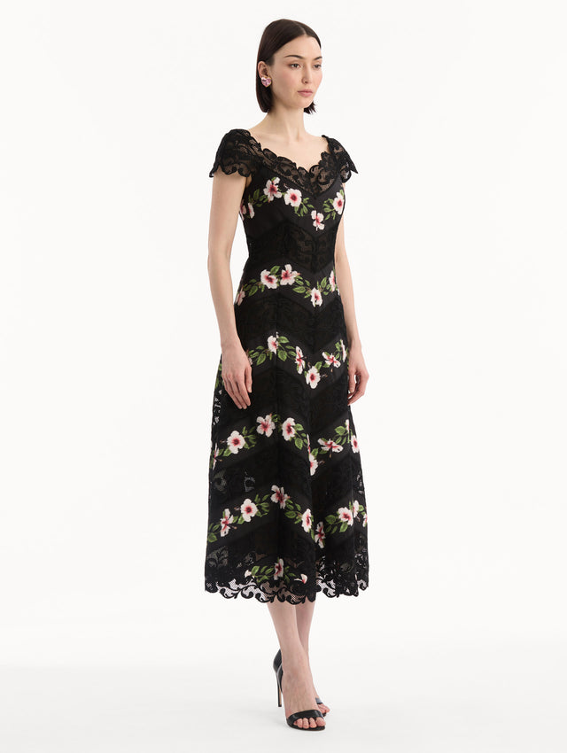 The model is wearing the Chiné Hibiscus Striped Faille Midi Dress in Black. Featuring off-the-shoulder cap sleeves, a midi length, and woven hibiscus motifs alternating with panels of faille and guipure lace, creating a striped composition. Side View.