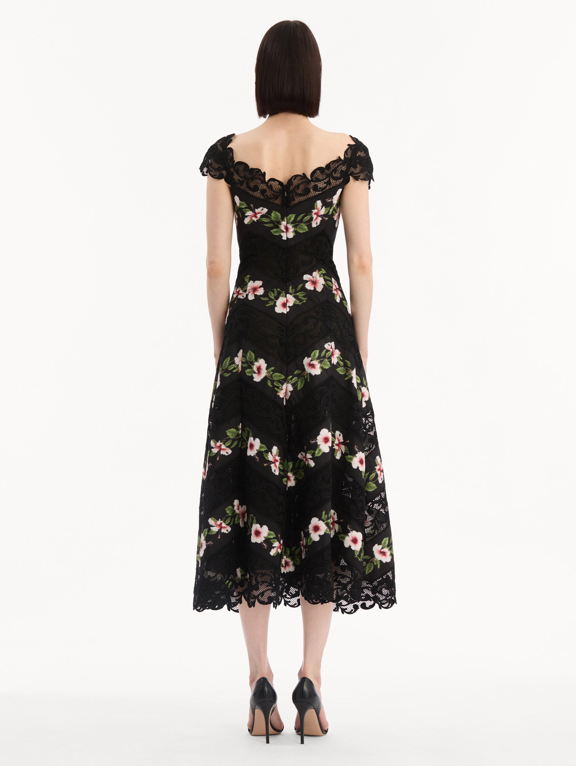 The model is wearing the Chiné Hibiscus Striped Faille Midi Dress in Black. Featuring off-the-shoulder cap sleeves, a midi length, and woven hibiscus motifs alternating with panels of faille and guipure lace, creating a striped composition. Back View.
