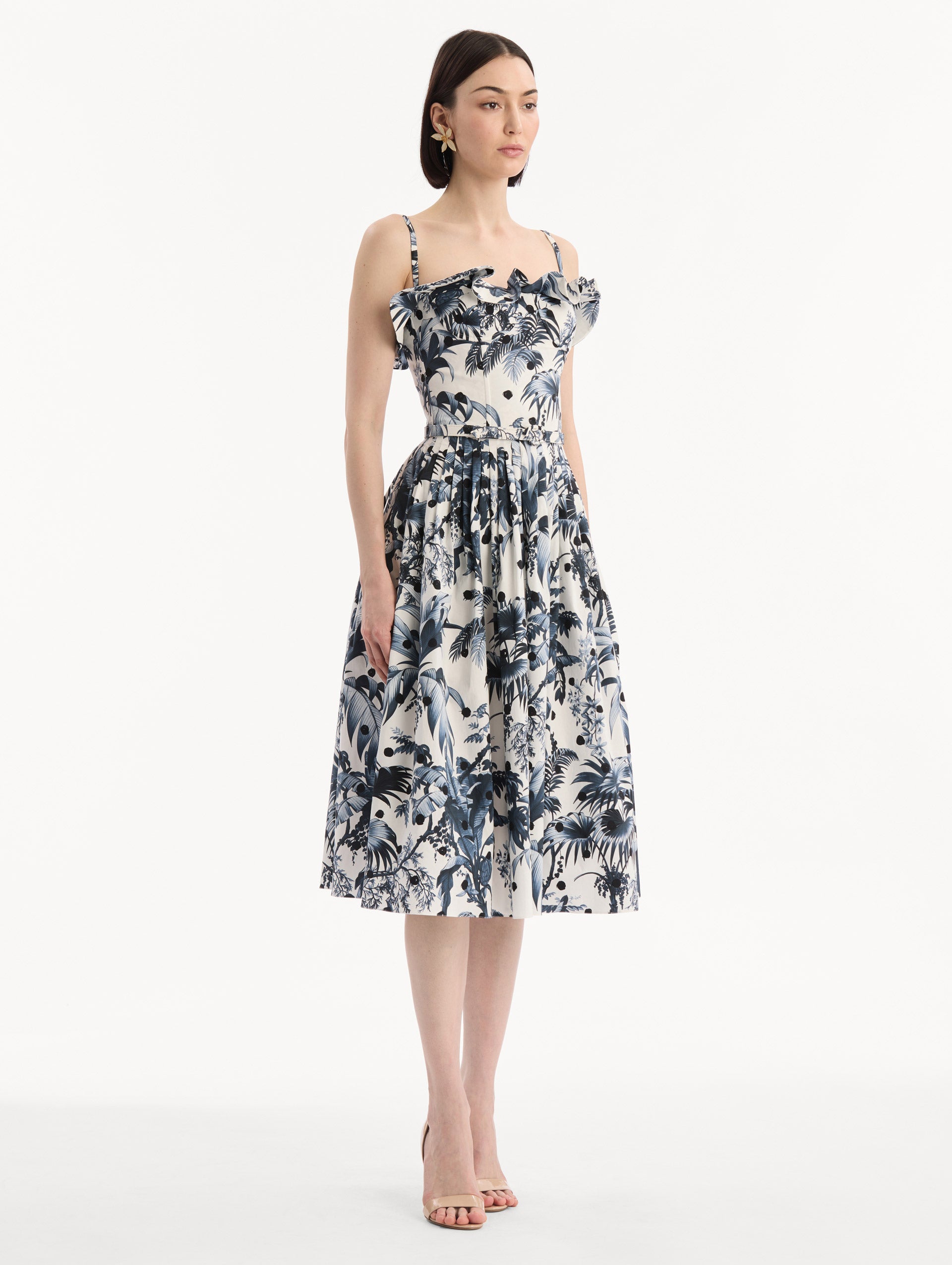 The model is wearing the Dotted Palm Leaves Cotton Poplin Sleeveless Dress in Navy Blue and White. Featuring a palm leaves print, ruffled top, delicate straps, belt at waist, softly structured bodice, and gathered skirt. Side View.