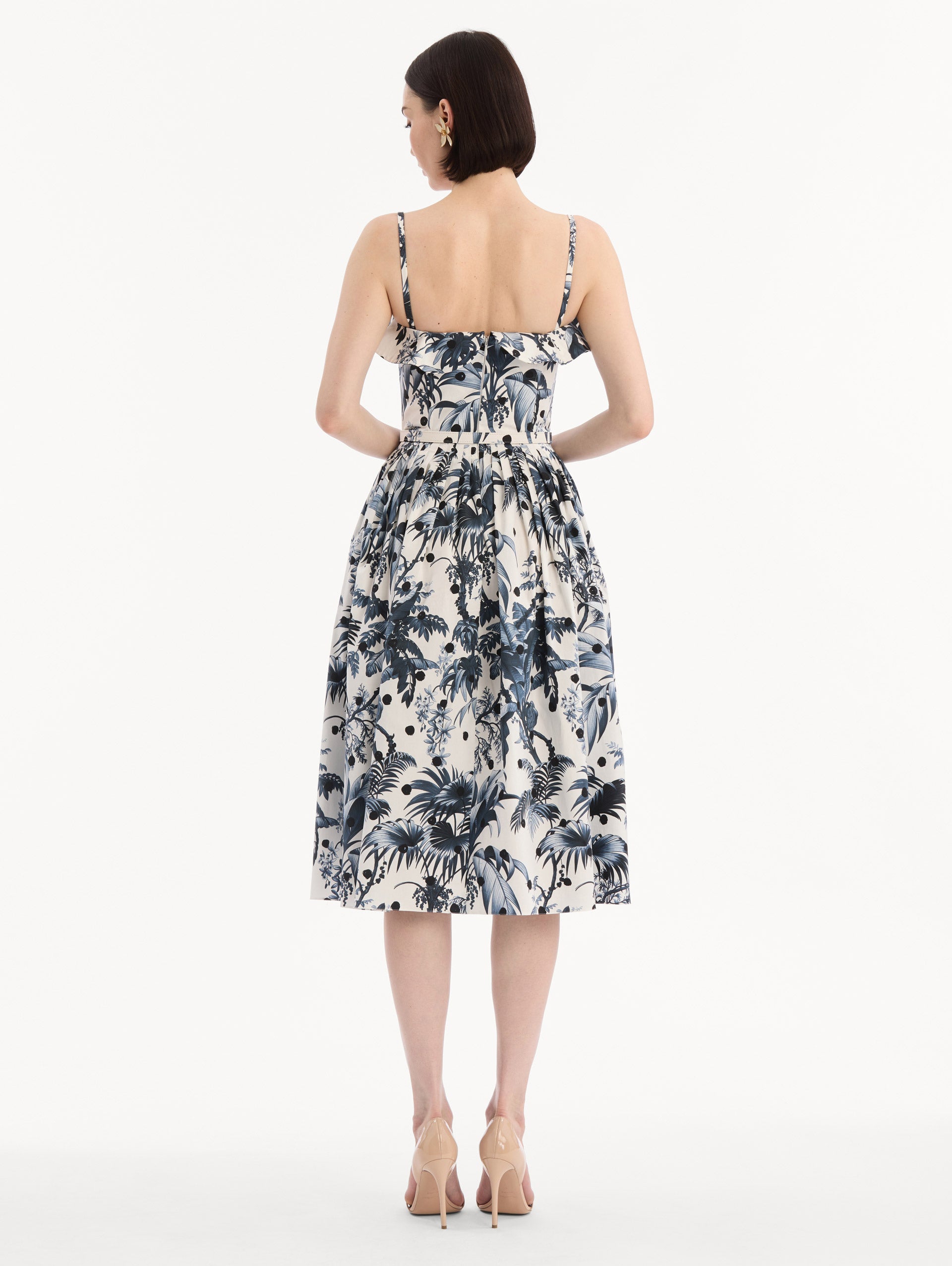 The model is wearing the Dotted Palm Leaves Cotton Poplin Sleeveless Dress in Navy Blue and White. Featuring a palm leaves print, ruffled top, delicate straps, belt at waist, softly structured bodice, and gathered skirt. Back View.