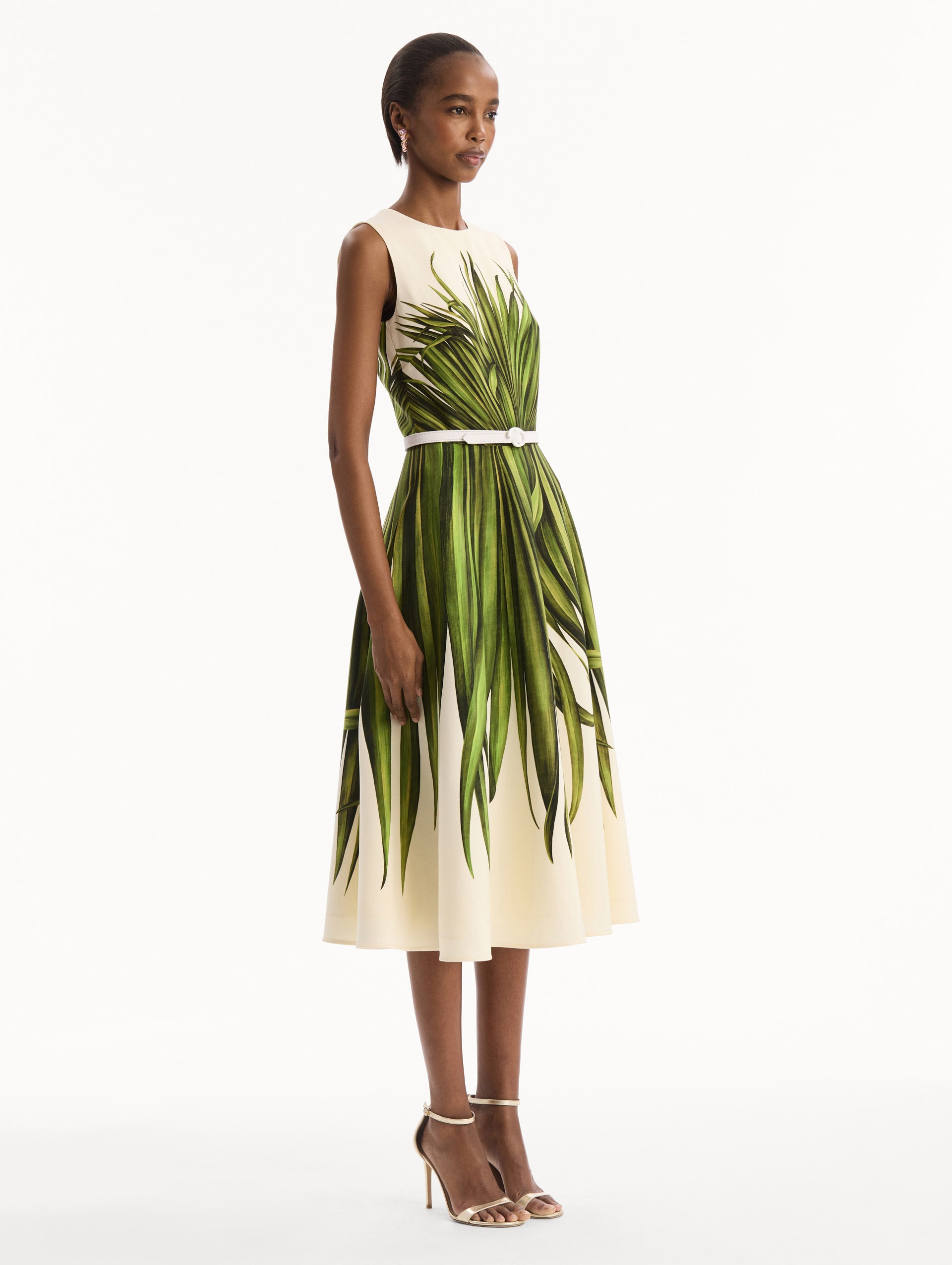 The model is wearing the Palm Leaves Wool-Blend Dress in ivory printed with green palm leaves. Featuring a belt at waist and midi-length skirt. Side View.