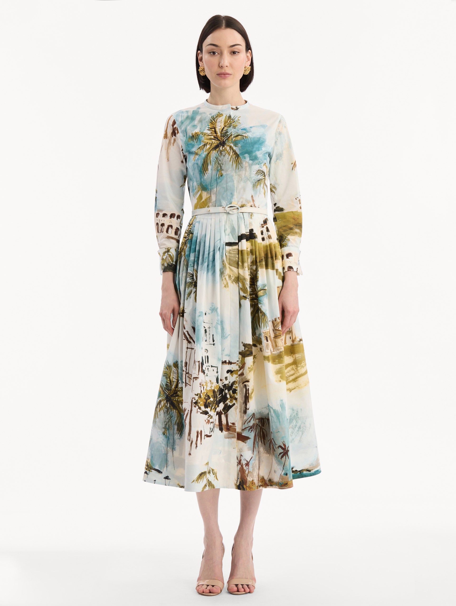 The model is wearing the Dominican Landscape Cotton Poplin Short-Sleeve Dress printed with the Dominican landscape. Featuring long sleeves, front-button closure, belt at waist, and pleated midi skirt. Front View.