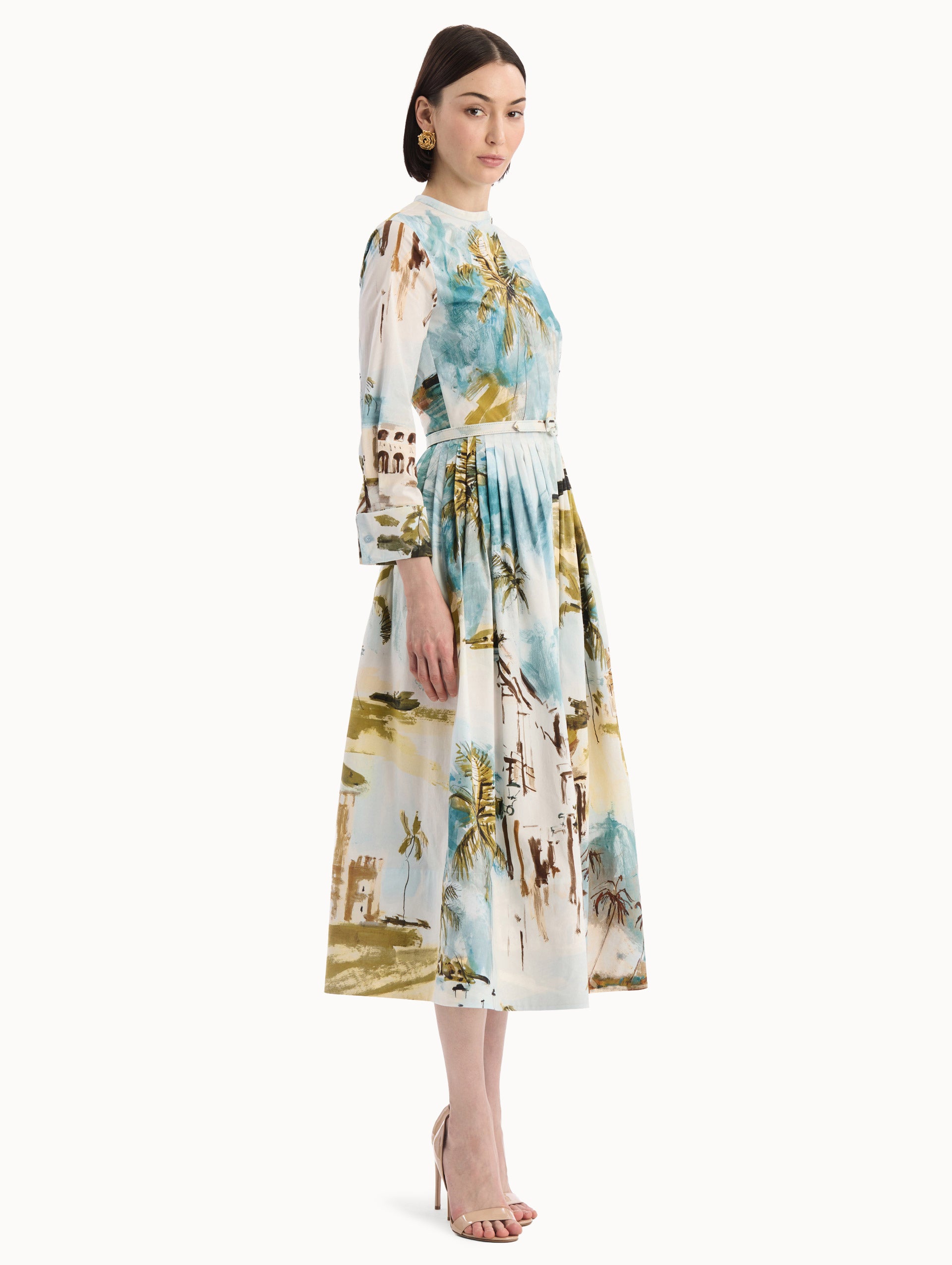 The model is wearing the Dominican Landscape Cotton Poplin Short-Sleeve Dress printed with the Dominican landscape. Featuring long sleeves, front-button closure, belt at waist, and pleated midi skirt. Side View.