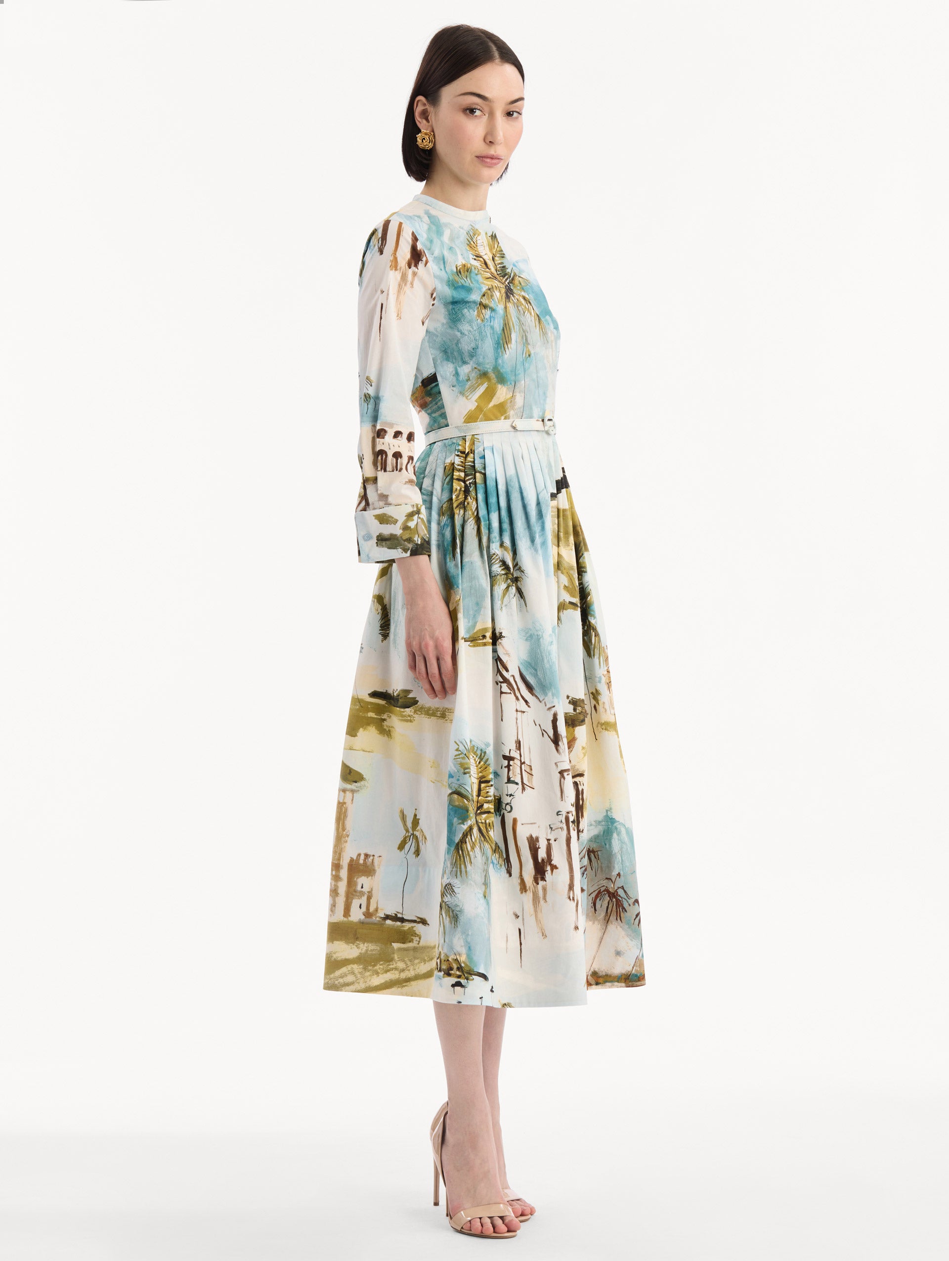 The model is wearing the Dominican Landscape Cotton Poplin Short-Sleeve Dress printed with the Dominican landscape. Featuring long sleeves, front-button closure, belt at waist, and pleated midi skirt. Side View.