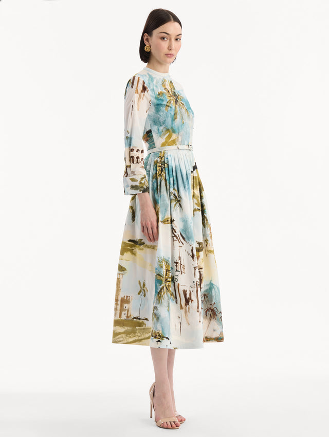 The model is wearing the Dominican Landscape Cotton Poplin Short-Sleeve Dress printed with the Dominican landscape. Featuring long sleeves, front-button closure, belt at waist, and pleated midi skirt. Side View.