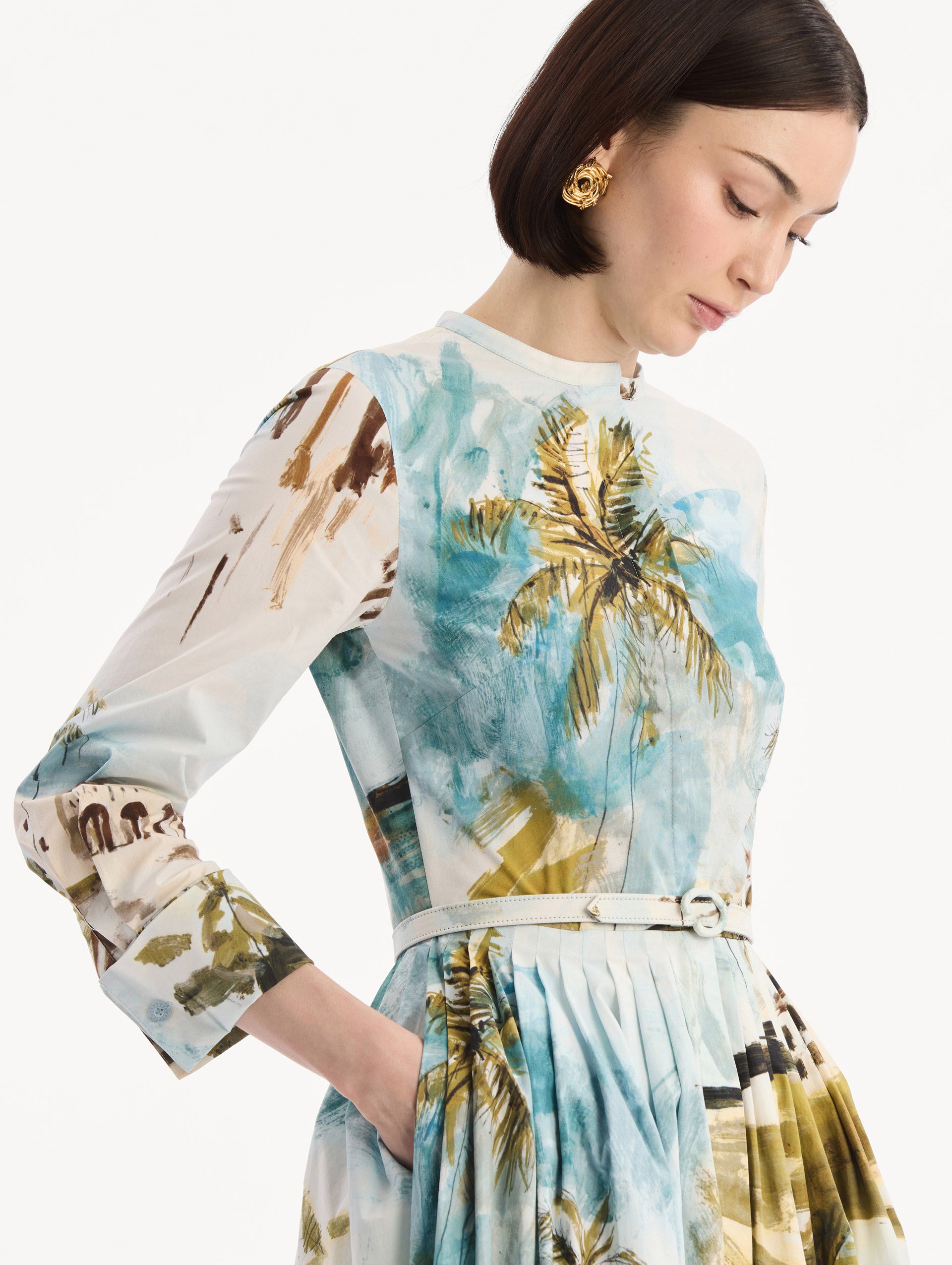 The model is wearing the Dominican Landscape Cotton Poplin Short-Sleeve Dress printed with the Dominican landscape. Featuring long sleeves, front-button closure, belt at waist, and pleated midi skirt. Detail View.