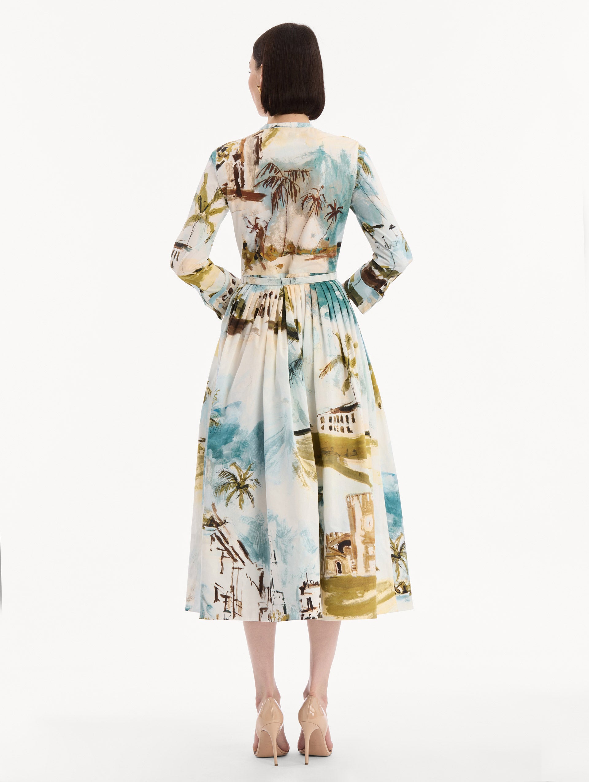 The model is wearing the Dominican Landscape Cotton Poplin Short-Sleeve Dress printed with the Dominican landscape. Featuring long sleeves, front-button closure, belt at waist, and pleated midi skirt. Back View.