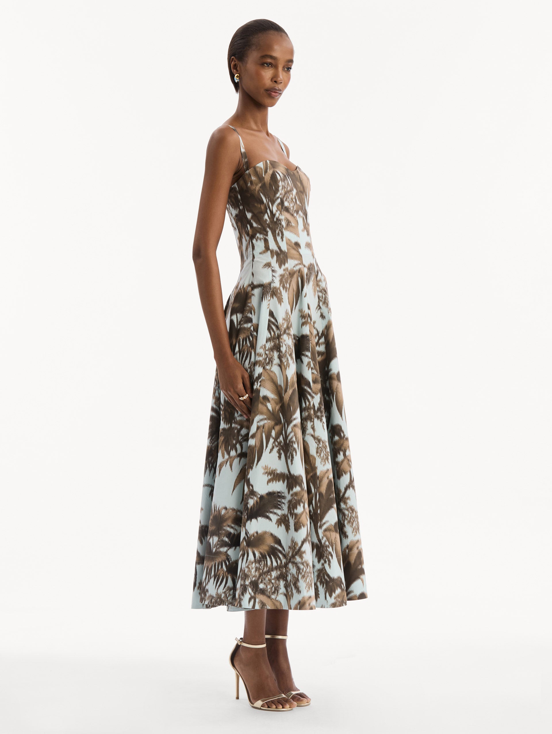 The model is wearing the Chiné Palm Leaves Cotton Poplin Dress in light blue printed with brown palm leaves. Featuring a subtle sweetheart neckline with tank straps, a softly structured bodice, and flowing midi skirt. Side View.